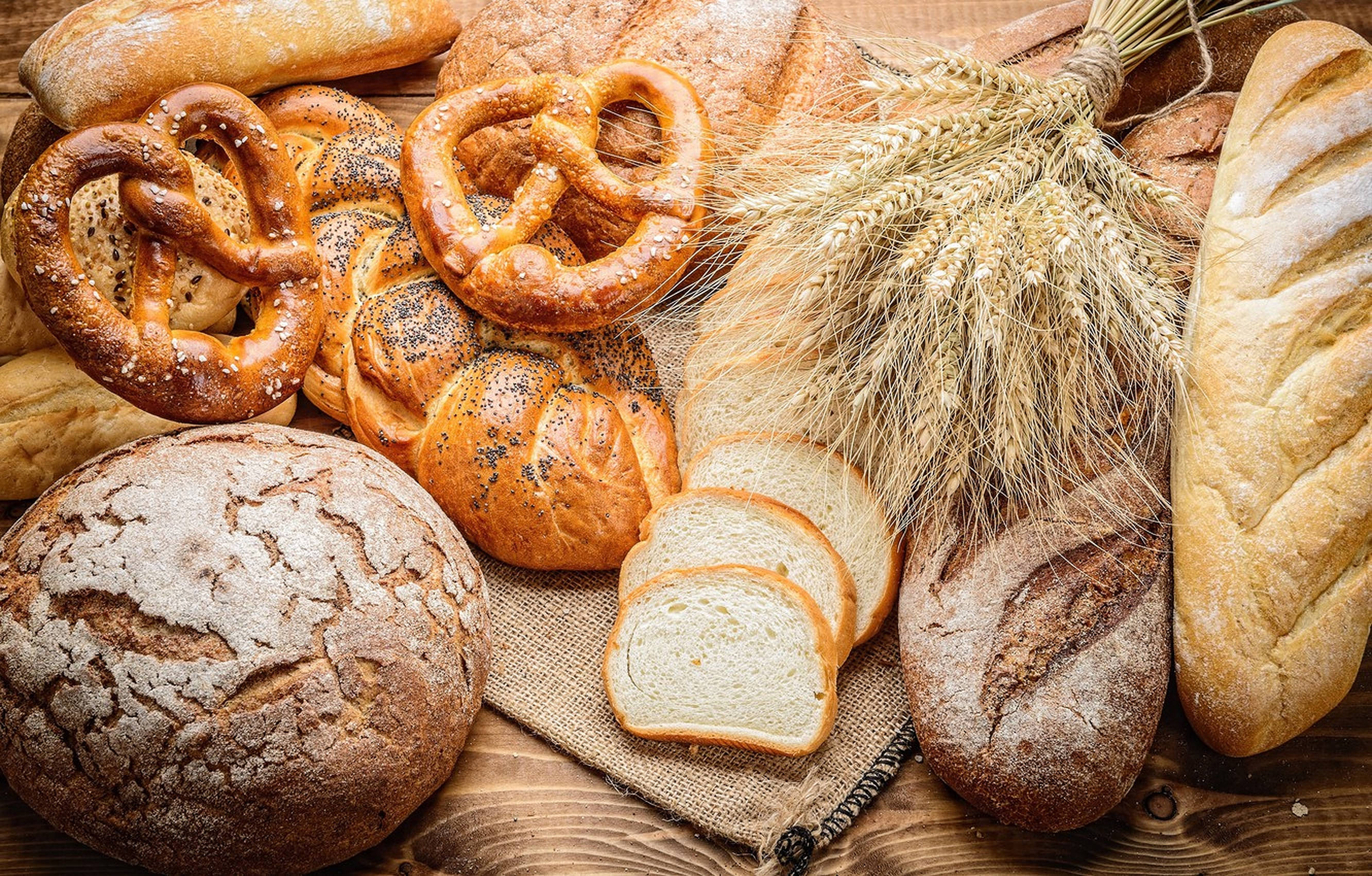 Bakery 桌布 壁紙 — 一張質樸的木桌上擺放著各種新鮮出爐的麵包、蝴蝶餅、麵包卷和小麥穗，搭配溫暖的金色燈光。