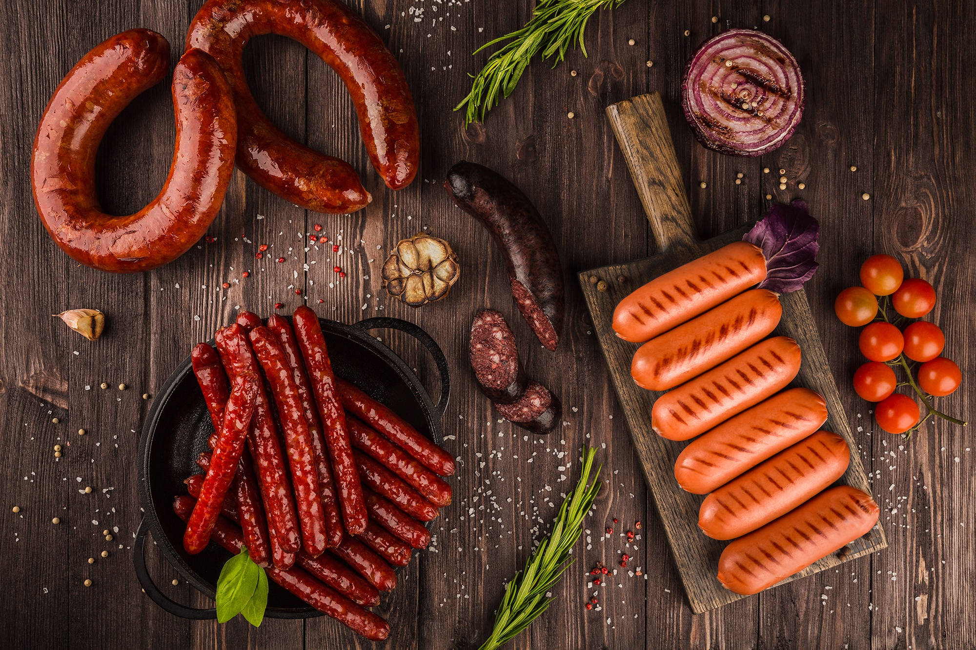 meat 桌布 壁紙 — 一張質樸的木桌上擺放著各種燒烤和生香腸，配以新鮮香草、櫻桃番茄和大蒜，營造出豐盛的烹飪場景。