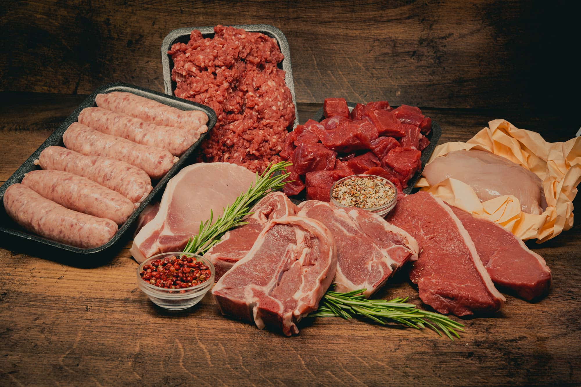 meat 桌布 壁紙 — 一張質樸的木桌上擺放著各種生肉，包括香腸、碎牛肉、牛排、豬排和雞胸肉，並以迷迭香和胡椒粒裝飾。