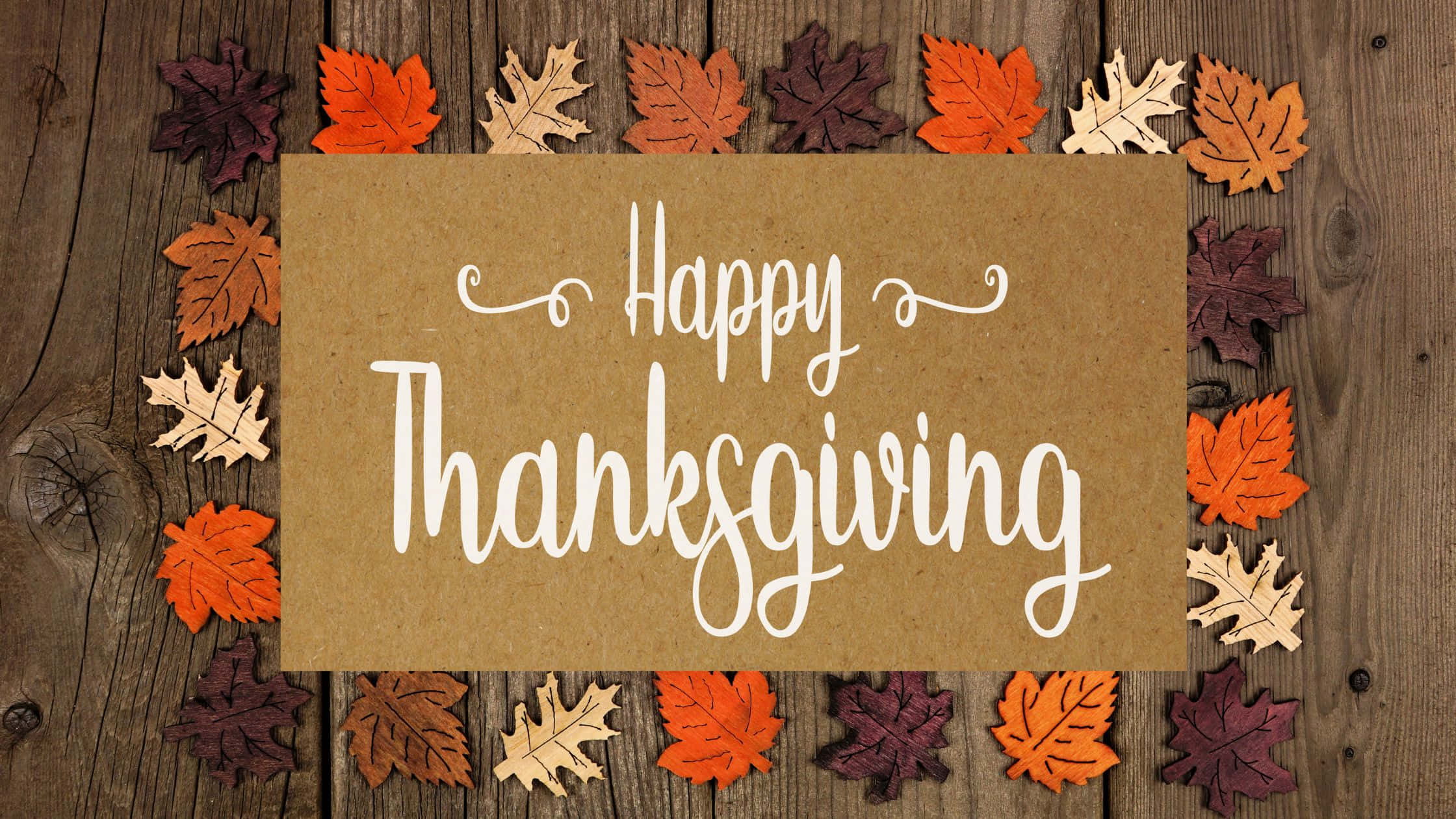 Thanksgiving Desktop 桌布 壁紙 — 一張質樸的棕色卡片，上面有白色草書「Happy Thanksgiving」文字，周圍環繞著色彩繽紛的秋葉，放置在風化的木質表面上。