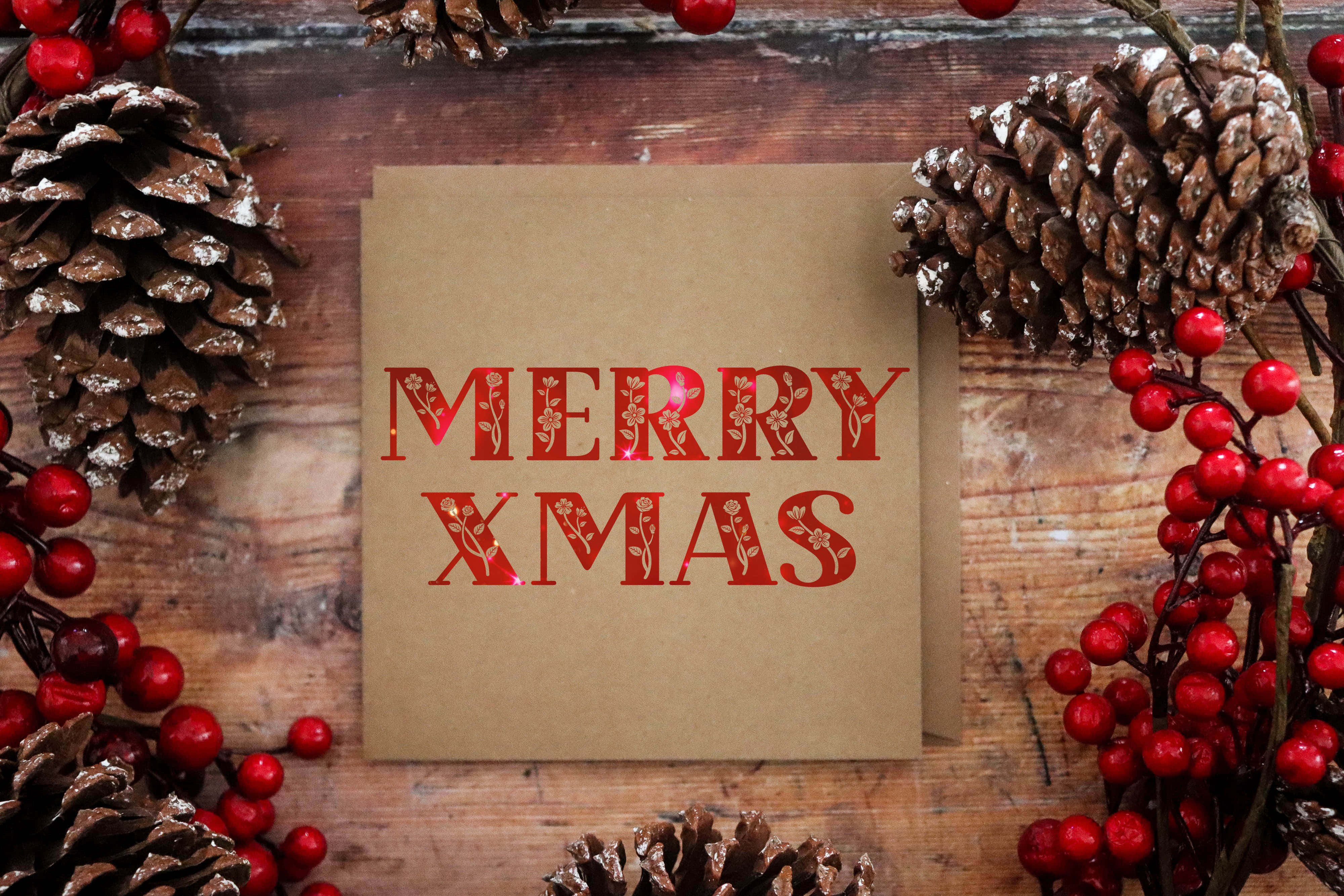 節日 桌布 壁紙 — 一張質樸的棕色卡片，上面有紅色的「MERRY XMAS」文字，周圍環繞著松果和紅色漿果，放在風化的木頭上。