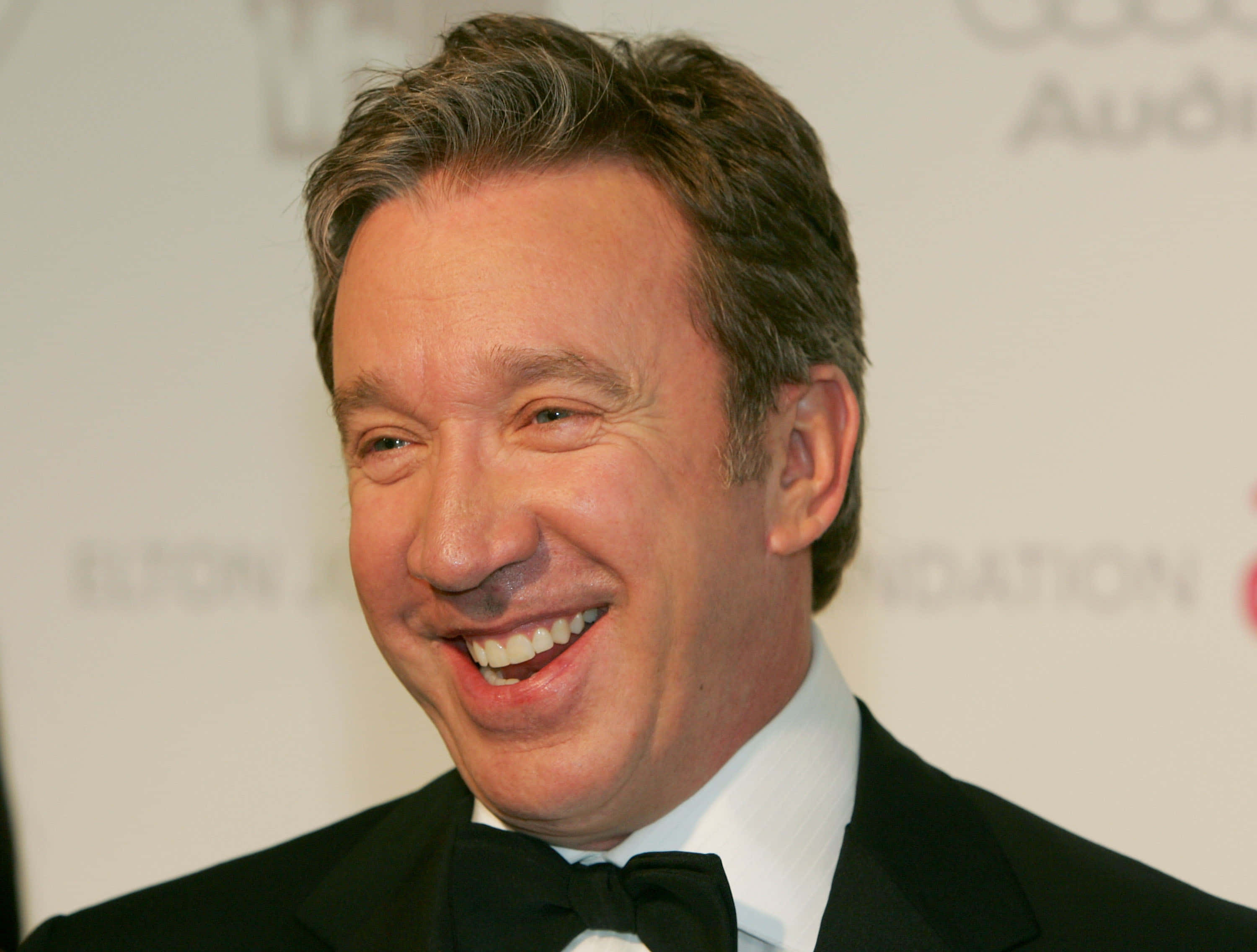 Tim Allen 桌布 壁紙 — 一張近距離照片，一位微笑的男子穿著黑色燕尾服和領結，在一個有品牌背景的正式活動中。