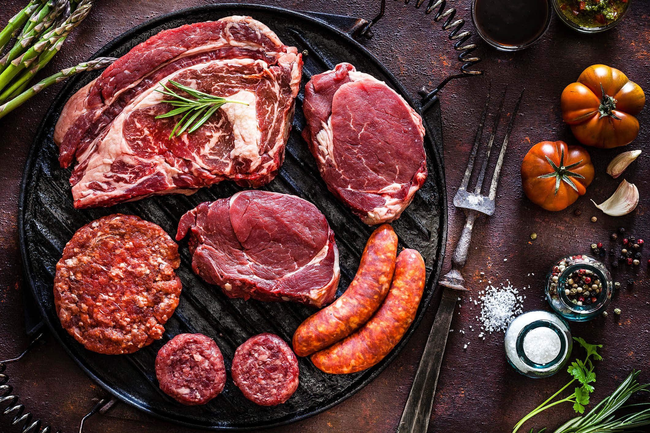 meat 桌布 壁紙 — 一張鄉村風格的俯拍照片，展示生肉，包括牛排、香腸和漢堡肉餅，排列在黑色烤盤上，周圍有新鮮香草和香料。