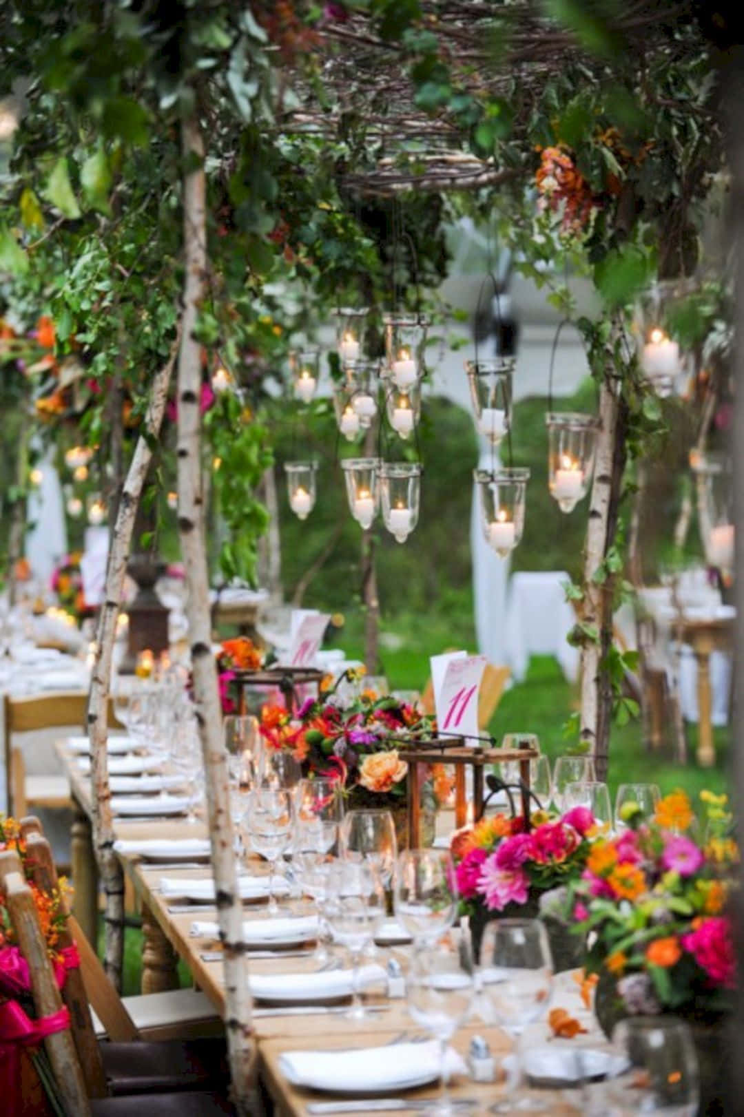 Garden Wedding 桌布 壁紙 — 一張長木桌，在枝葉茂密的涼亭下為花園婚禮佈置，掛有蠟燭和鮮豔的花卉中心裝飾。