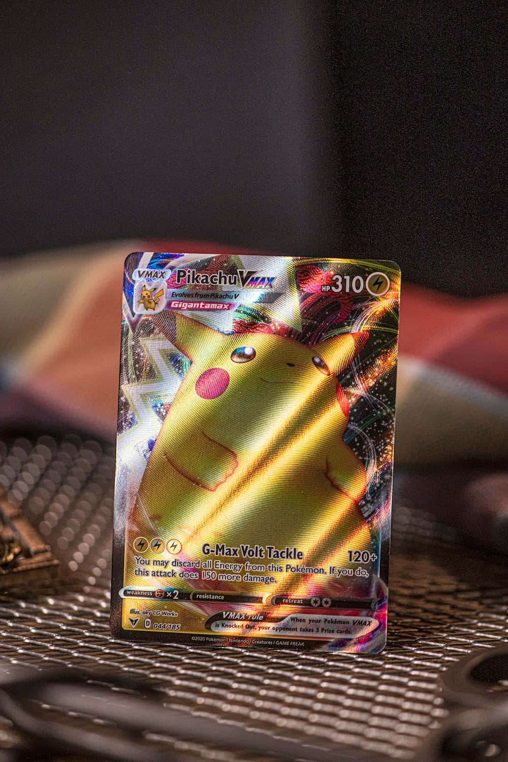 Pokemon Card 桌布 壁紙 — 一張閃亮的皮卡丘VMAX Pokémon卡，帶有全息金色和彩虹箔片細節，放置在紋理金屬表面上。