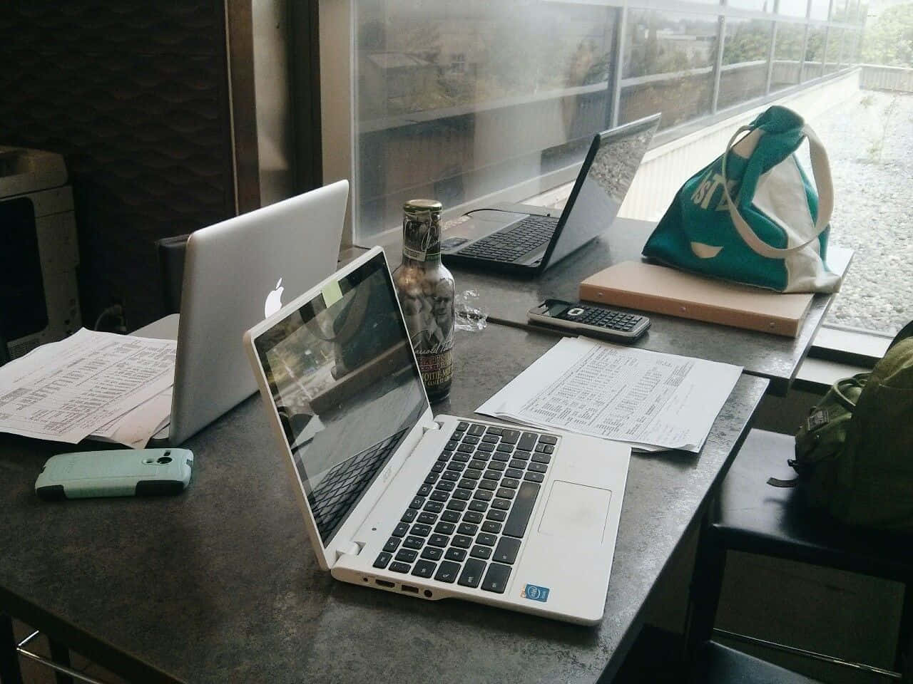 Studying 桌布 壁紙 — 一張雜亂的書桌，上面有兩部打開的筆記型電腦、文件、一個瓶子和一個袋子，靠近一扇顯示下雨戶外場景的窗戶。