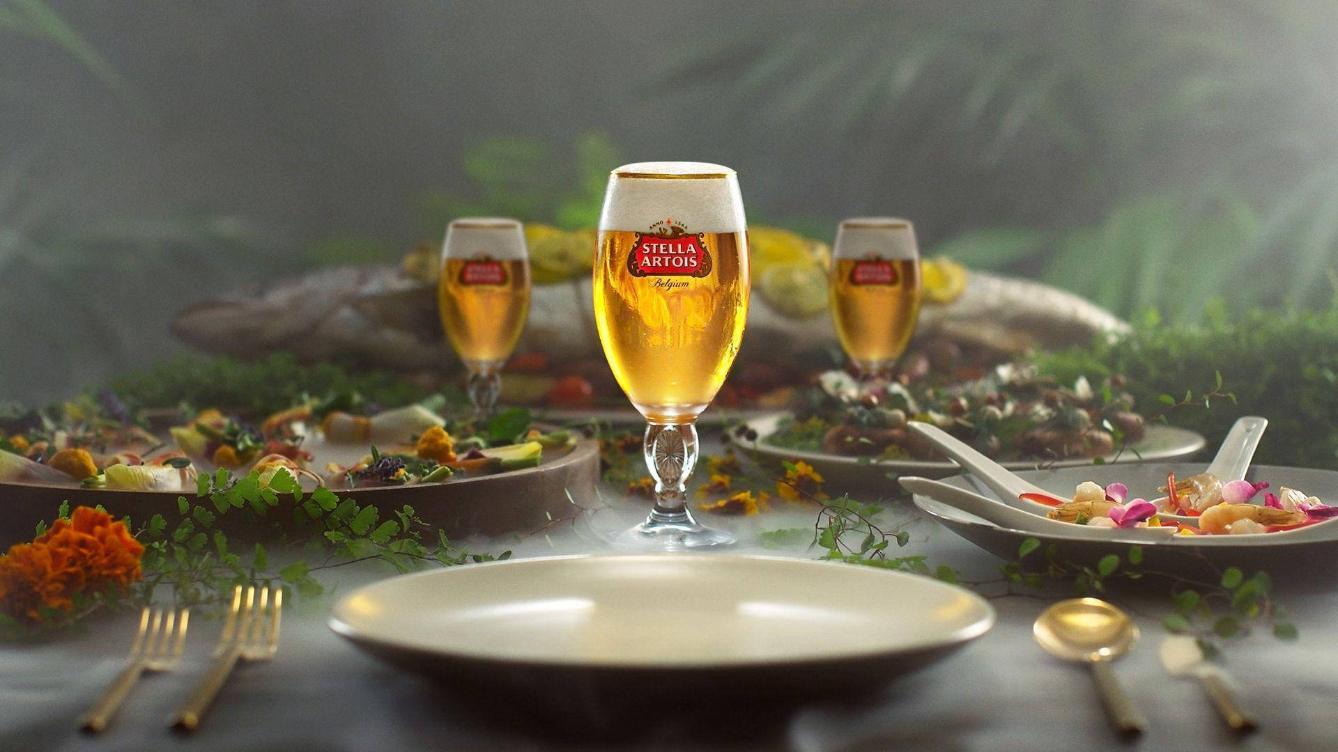 Beer 桌布 壁紙 — 一張霧氣瀰漫的桌子，擺放著 Stella Artois 啤酒和美食菜餚，周圍環繞著茂密的綠植。