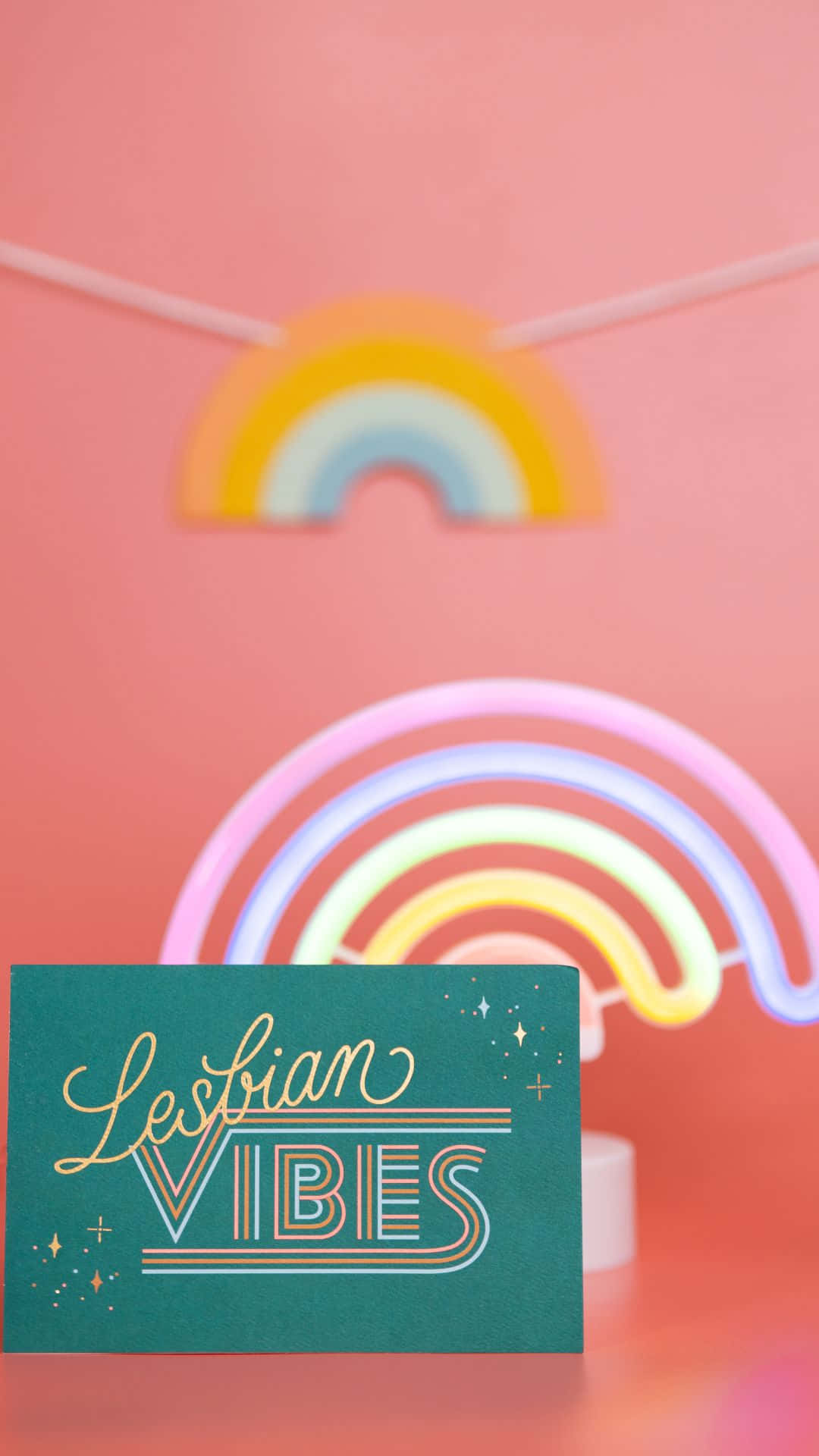 女同志 桌布 壁紙 — 一張青綠色卡片，上面寫著「Lesbian Vibes」，放在發光的霓虹彩虹前，背景是柔和的粉紅色牆壁。