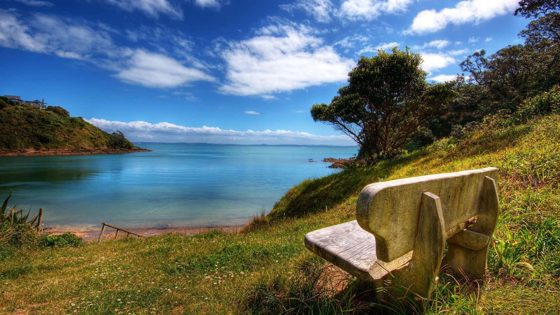 Relaxing Desktop 桌布 壁紙 — 一張風化的木凳放在長滿草的山丘上，俯瞰著平靜的碧綠海灣，天空湛藍，白雲朵朵。