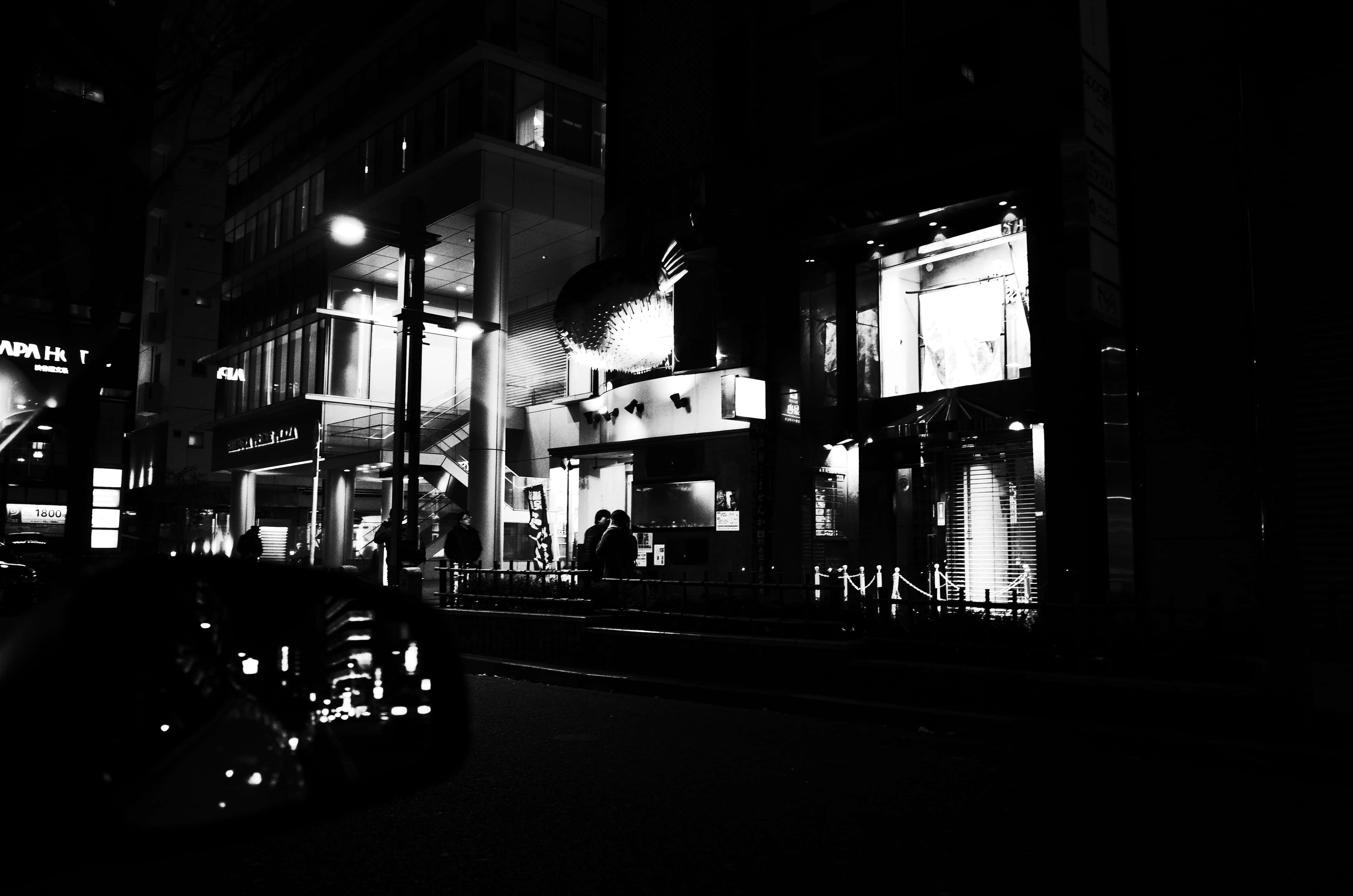 日本黑白 桌布 壁紙 — 一張高對比度的黑白城市街道夜景照片，有燈火通明的商店門面和剪影般的行人。