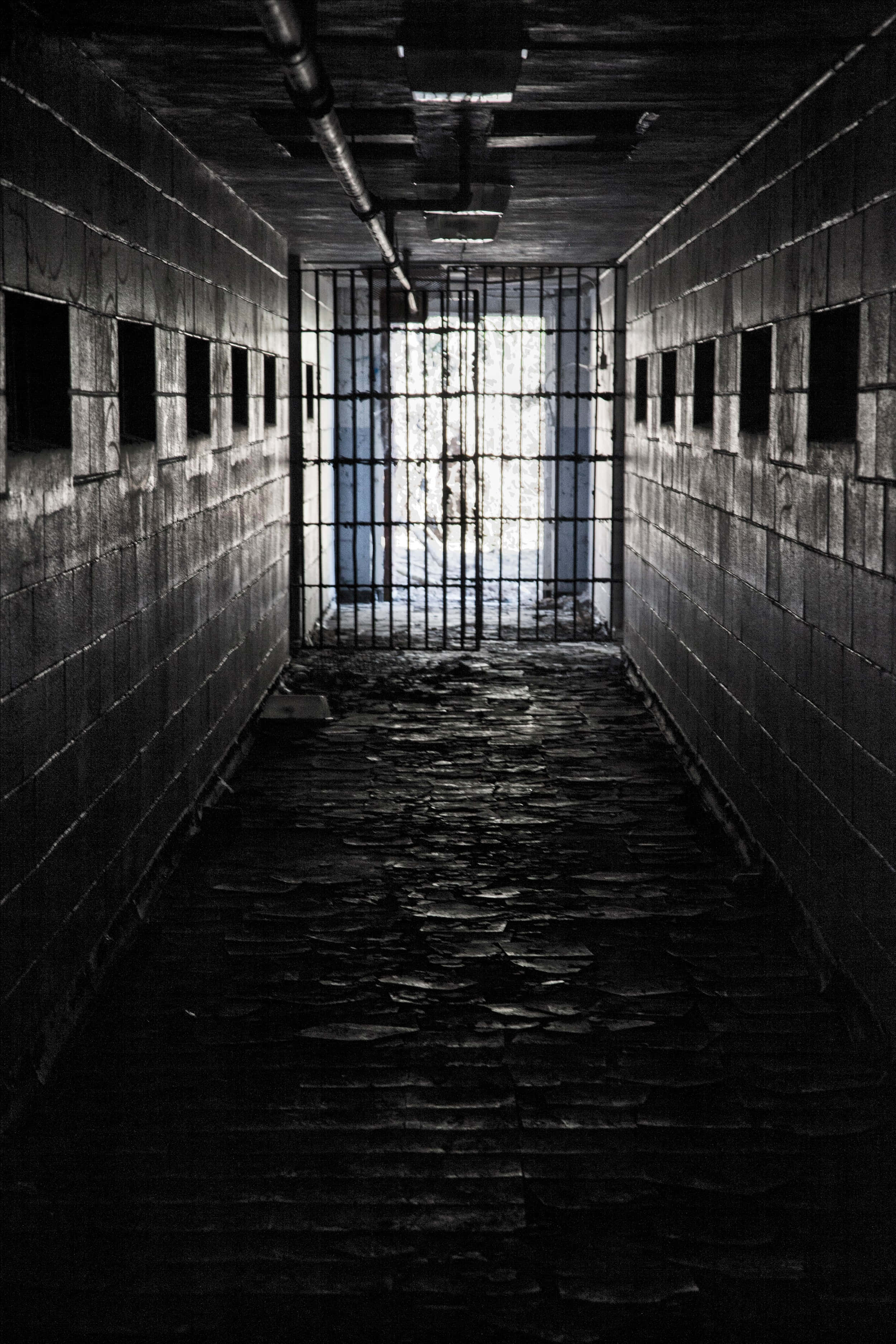 Jail 桌布 壁紙 — 一張鮮明的黑白照片，展示一條破舊的監獄走廊，有鐵欄門和破損的地磚，通向一道鐵欄閘門。