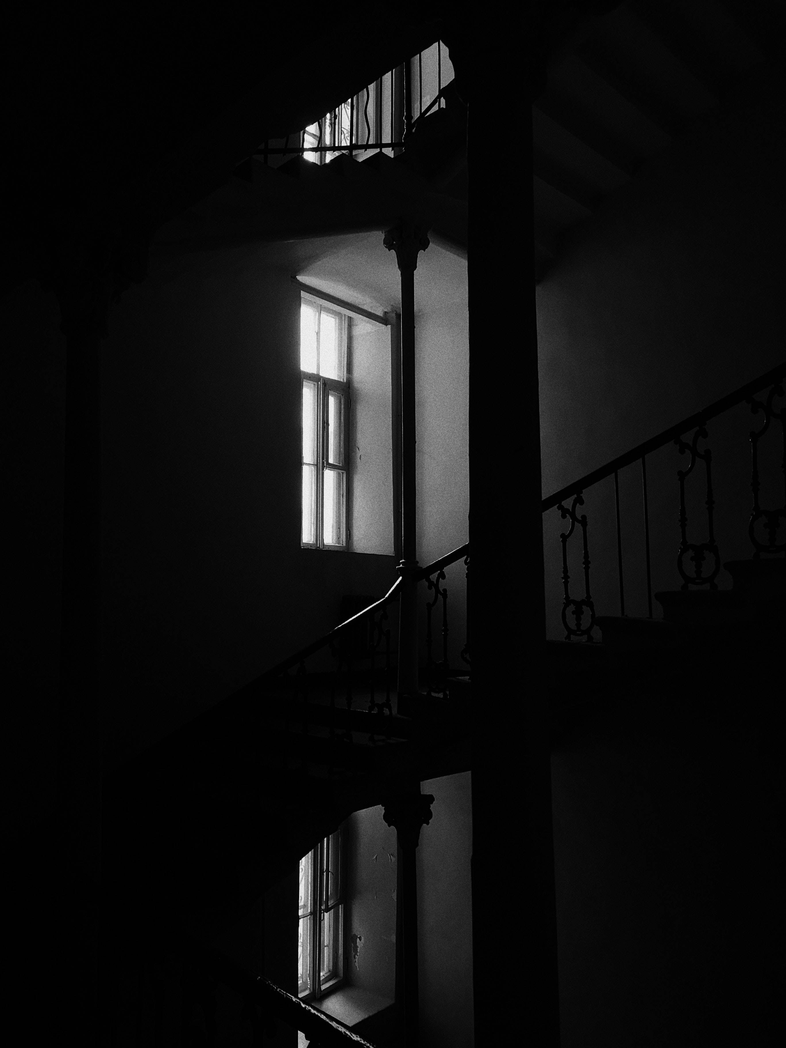 Dark Minimalist 桌布 壁紙 — 一張鮮明的黑白照片，展示光線昏暗的螺旋樓梯，帶有華麗的欄杆，由一扇窗戶照亮，投射出戲劇性的陰影。