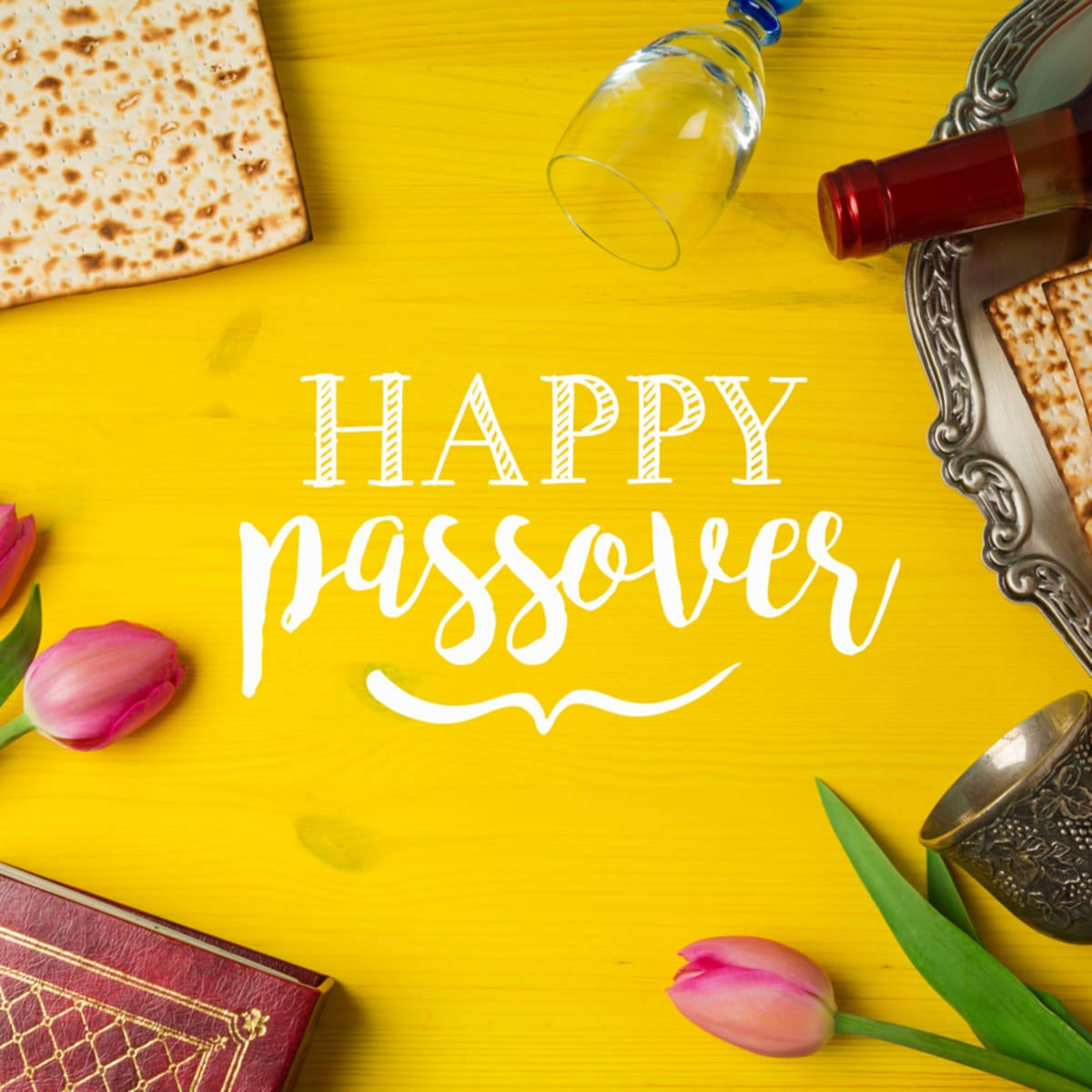 Passover 桌布 壁紙 — 一張鮮黃色木桌上展示逾越節象徵，包括無酵餅、酒杯和粉紅色鬱金香，並有「Happy Passover」字樣。