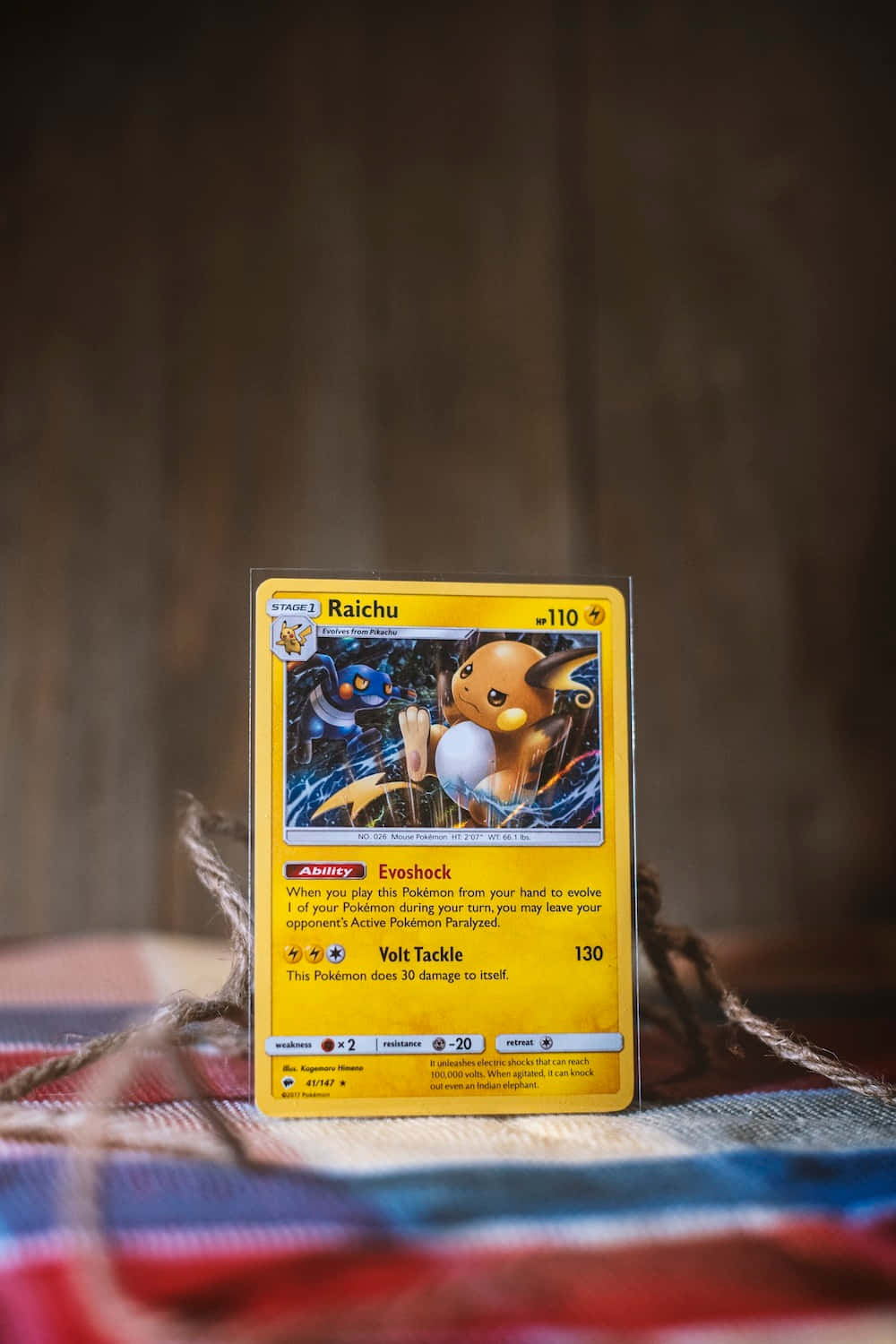 Pokemon Card 桌布 壁紙 — 一張黃色雷丘寶可夢卡直立於色彩繽紛的編織毯上，背景為模糊的木質。