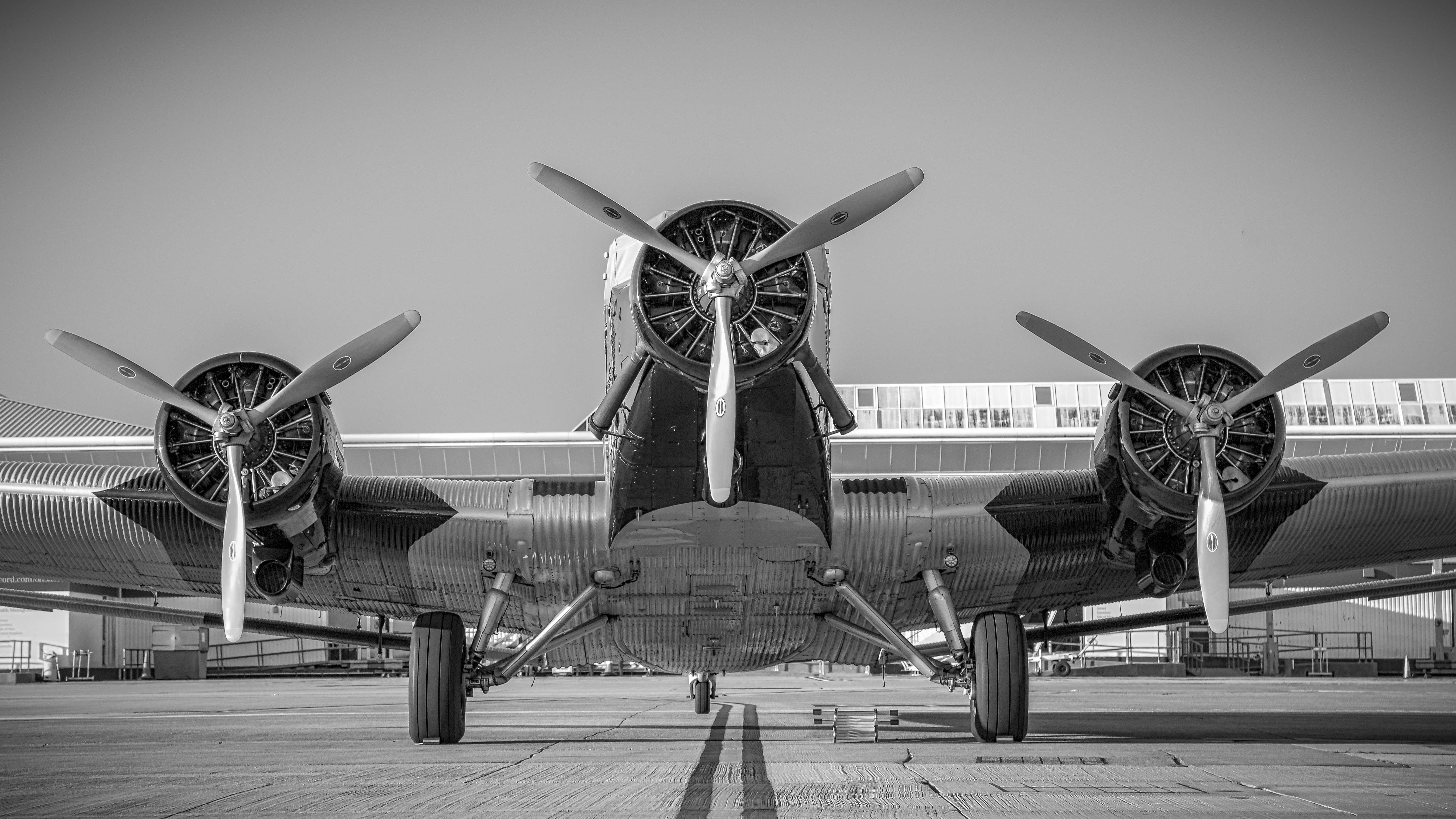Plane 桌布 壁紙 — 一張黑白低角度拍攝的復古三引擎螺旋槳飛機停在機場停機坪上的照片。