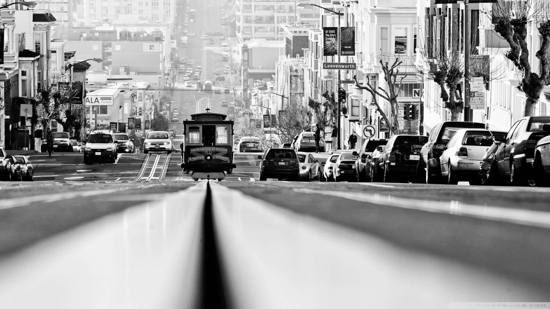 San Francisco Black And White 桌布 壁紙 — 一張黑白低角度拍攝的纜車照片，纜車正沿著舊金山一條陡峭的街道上行，街道兩旁停滿汽車和建築物。