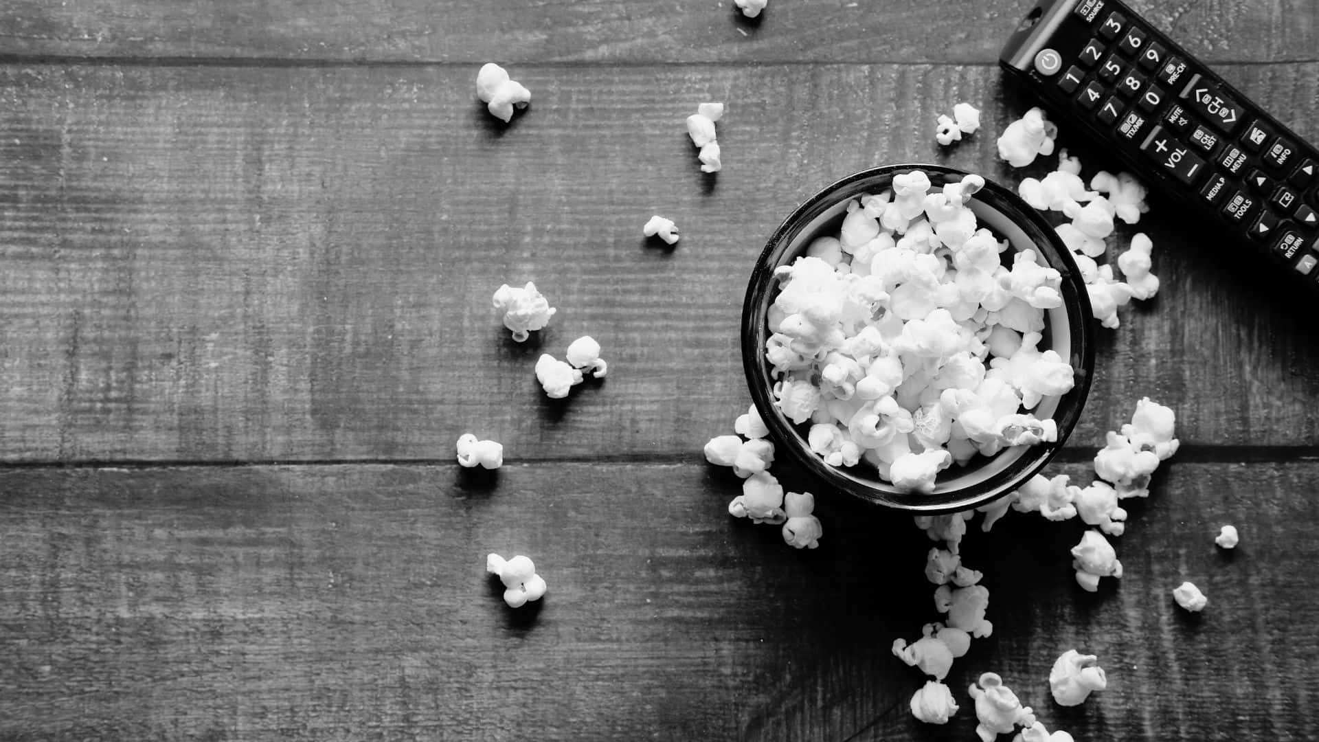 Black And White Movie 桌布 壁紙 — 一張黑白俯拍照片，顯示一碗爆米花灑落在深色木桌上，旁邊有一個遙控器。