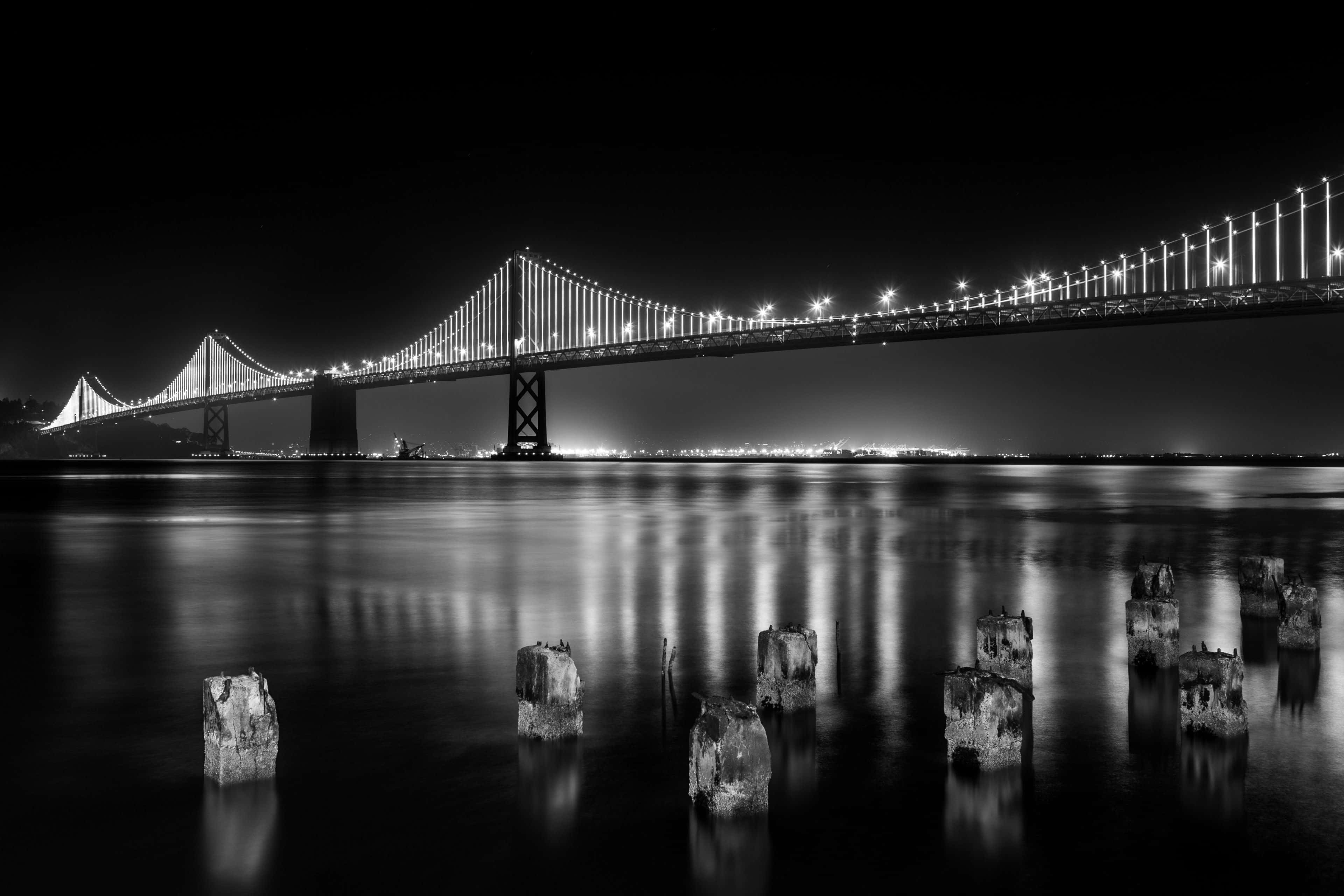 Black And White Architecture 桌布 壁紙 — 一張黑白夜景照片，拍攝了燈光璀璨的吊橋倒映在平靜的水面上，前景有風化的木樁。
