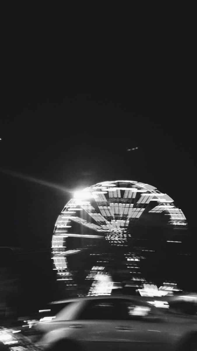 桌布 壁紙 — 一張黑白夜景照片，發光的摩天輪倒映在水中，一輛模糊的汽車駛過。