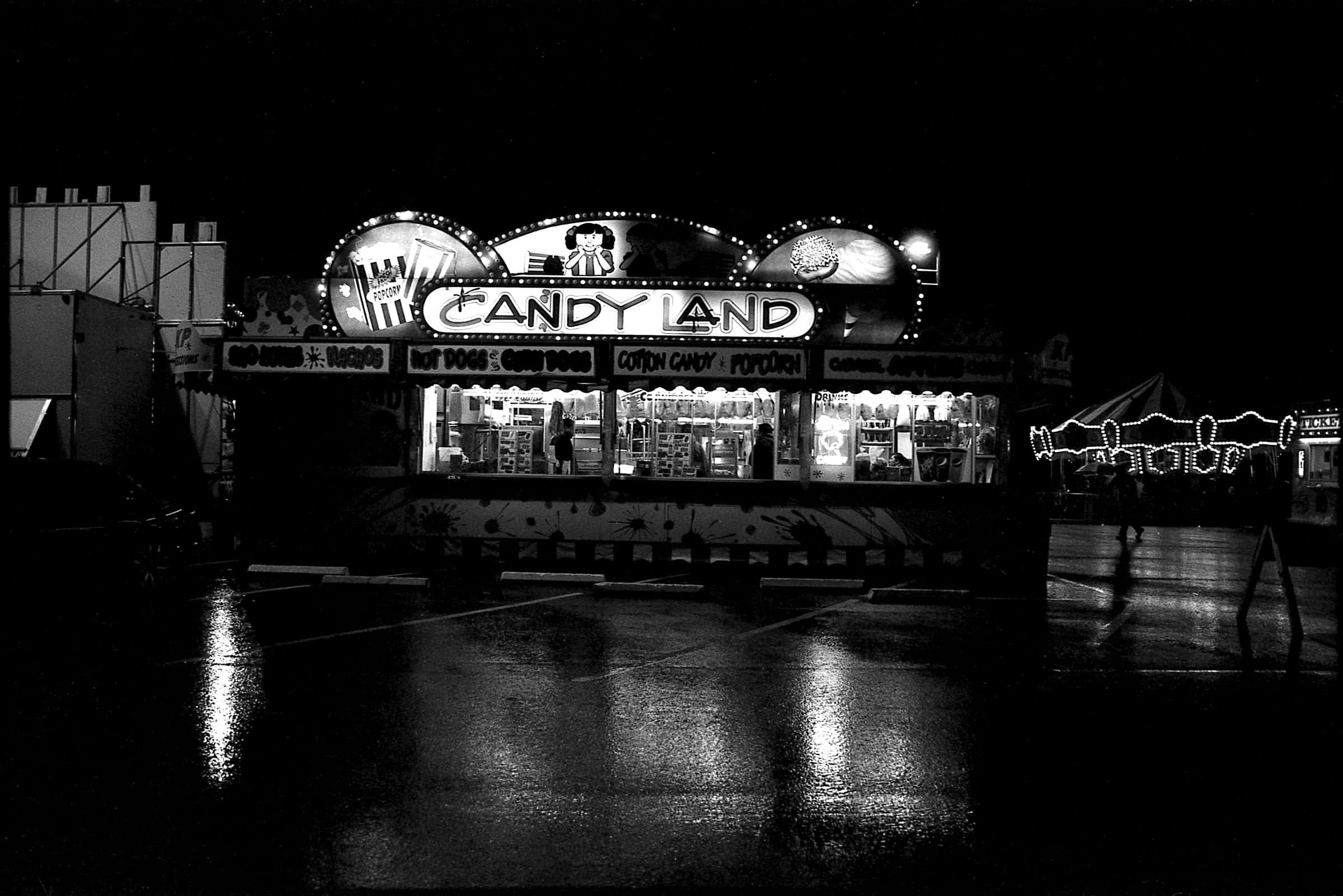 Dark Carnival 桌布 壁紙 — 一張黑白夜景照片，糖果攤在雨濕的路面上發光，帶有倒影和剪影。