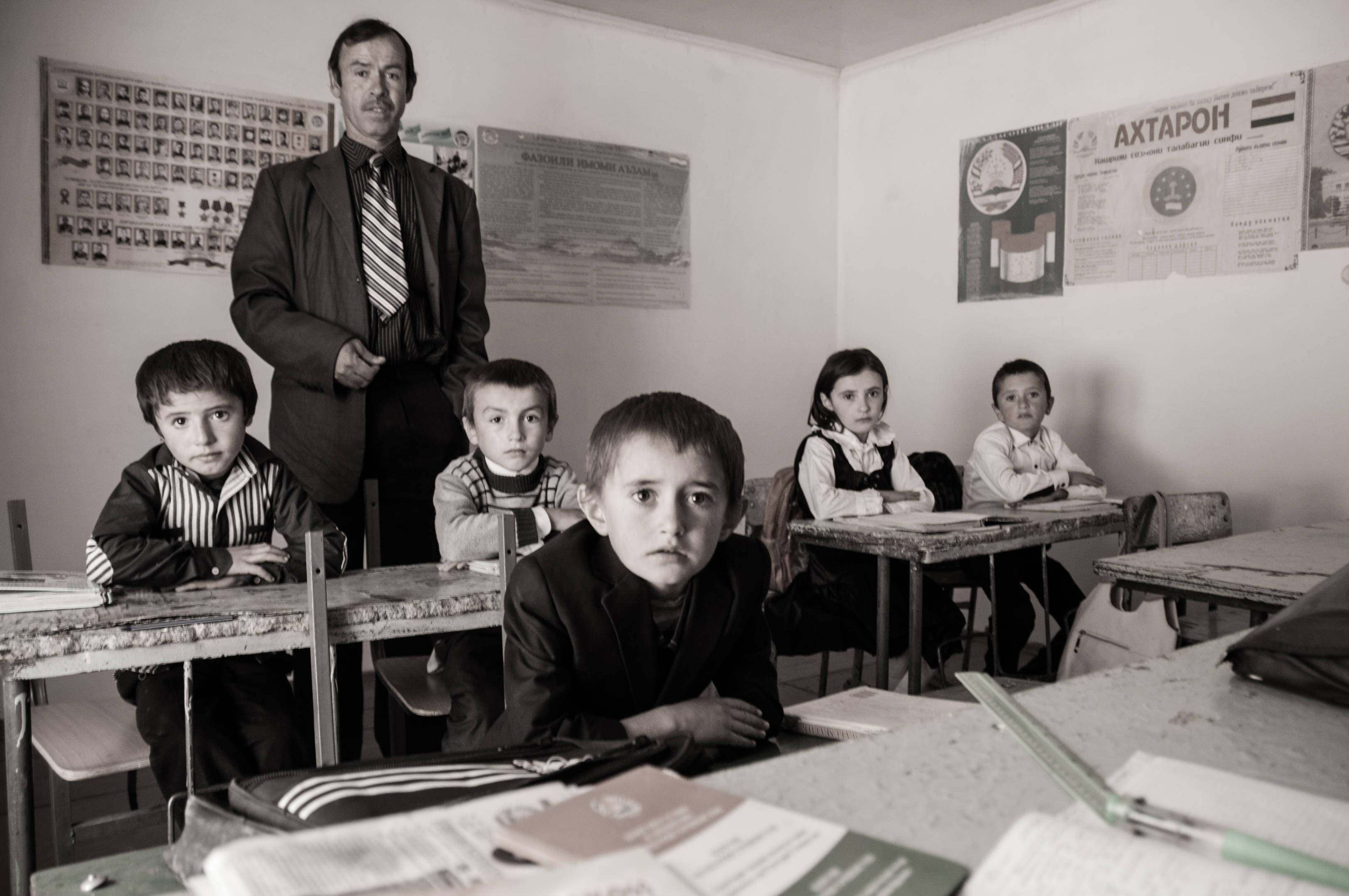 Tajikistan 桌布 壁紙 — 一張黑白照片，一位嚴肅的老師站在專注的孩子們身後，教室樸素，牆上掛著教育海報。
