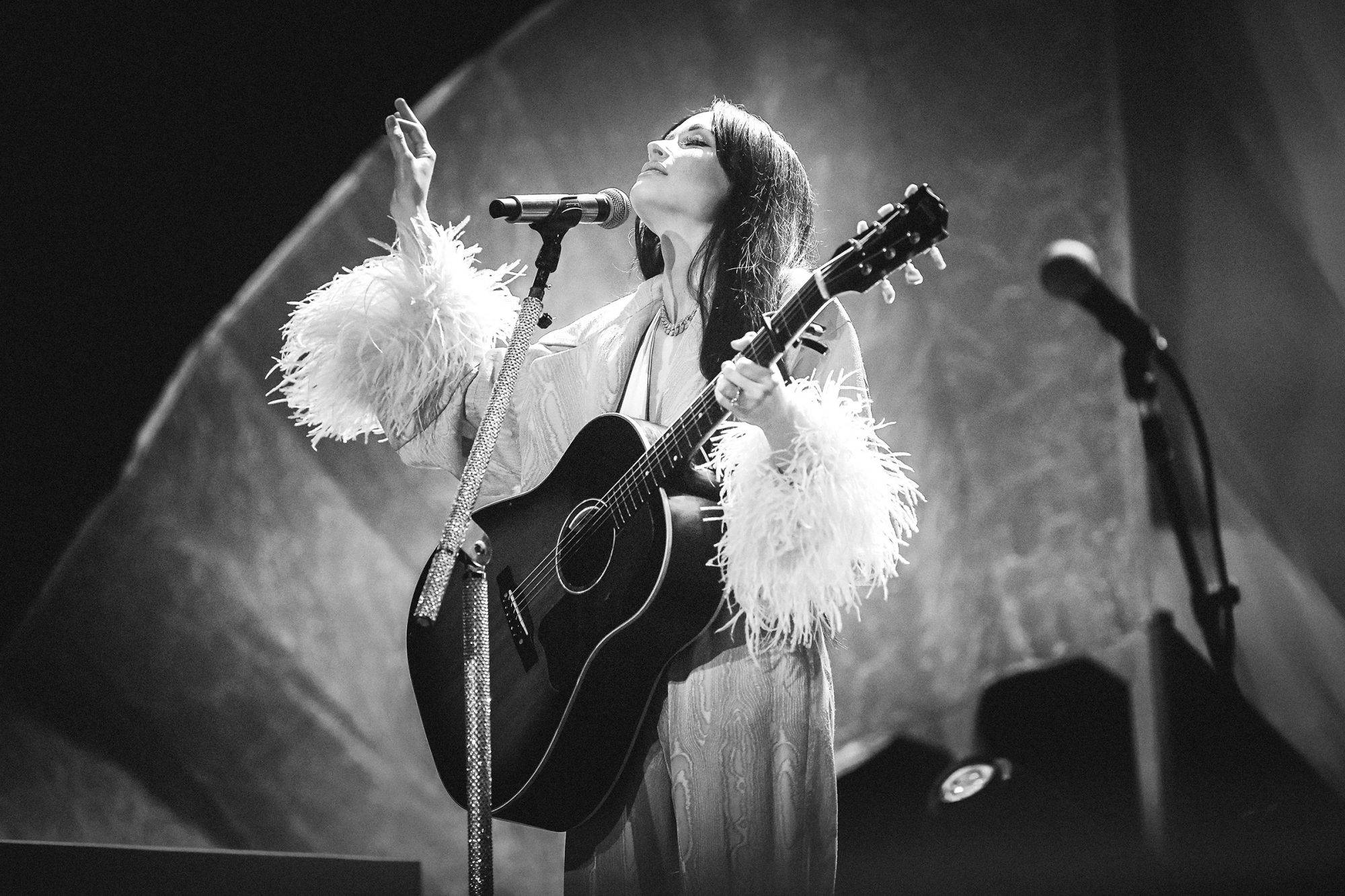 Kacey Musgraves 桌布 壁紙 — 一張黑白照片，一位女歌手穿著羽毛袖，在戲劇性燈光下熱情地彈奏吉他。