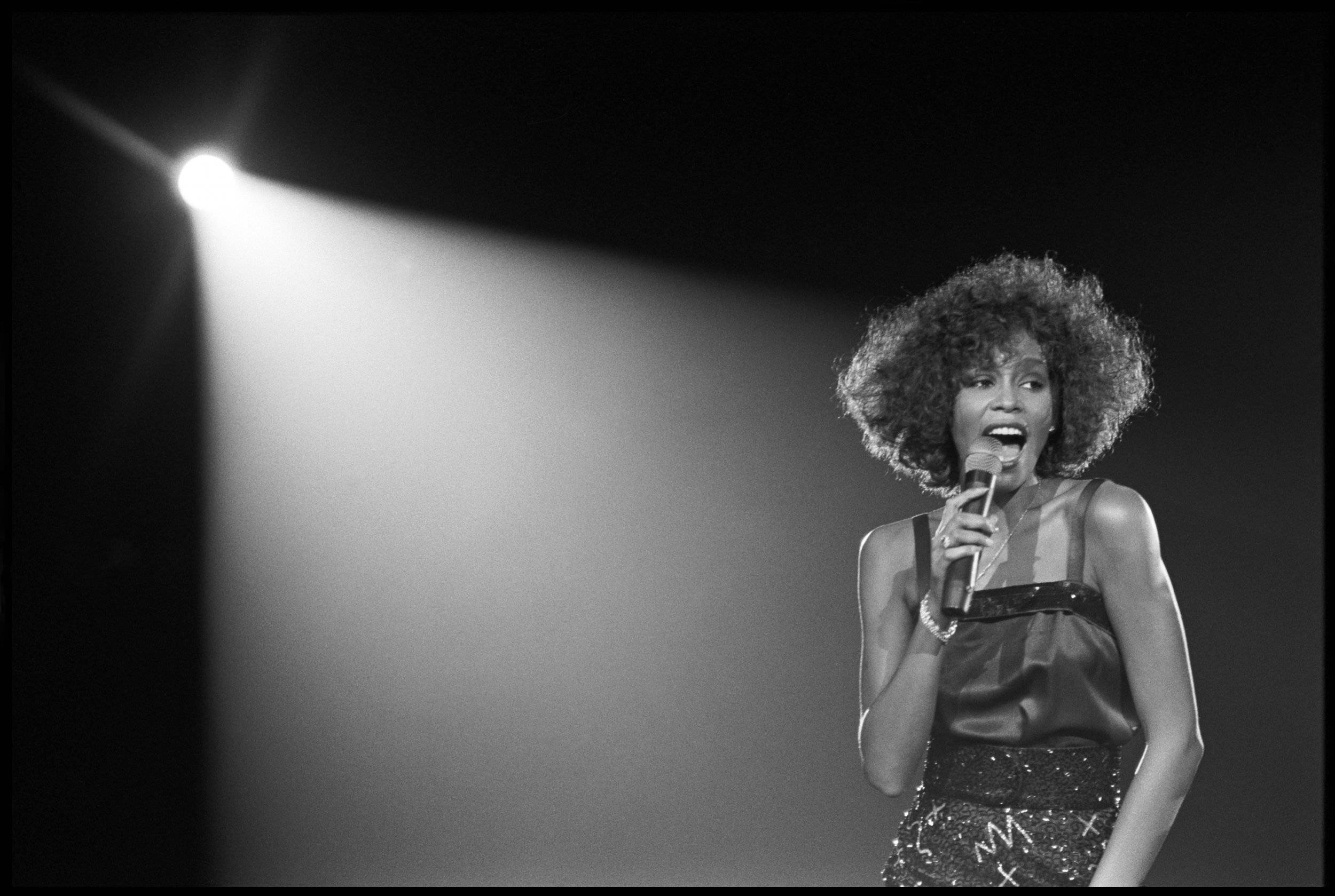 Whitney 桌布 壁紙 — 一張黑白照片，一位擁有豐滿爆炸頭的歌手在舞台上單一聚光燈下表演。