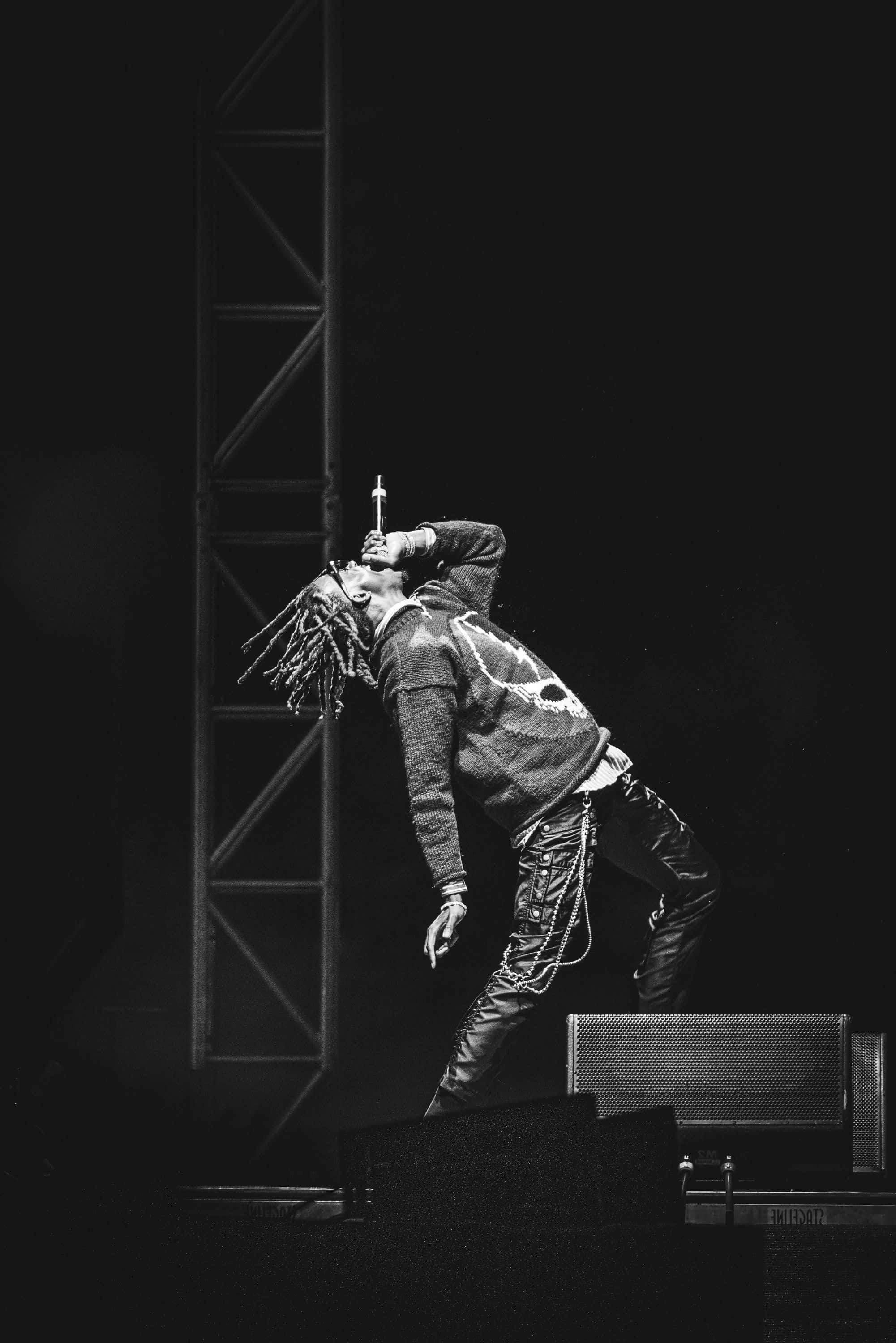Playboi Carti HD 桌布 壁紙 — 一張黑白照片，一位留著辮子的表演者在昏暗的舞台上熱情歌唱。