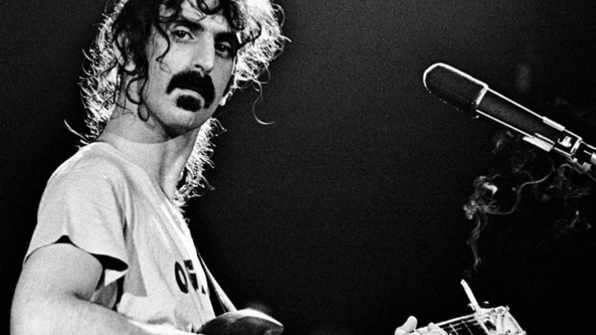 Zappa 桌布 壁紙 — 一張黑白照片，一位留鬍子、捲髮的音樂家在舞台上靠近麥克風彈吉他，煙霧裊裊升起。