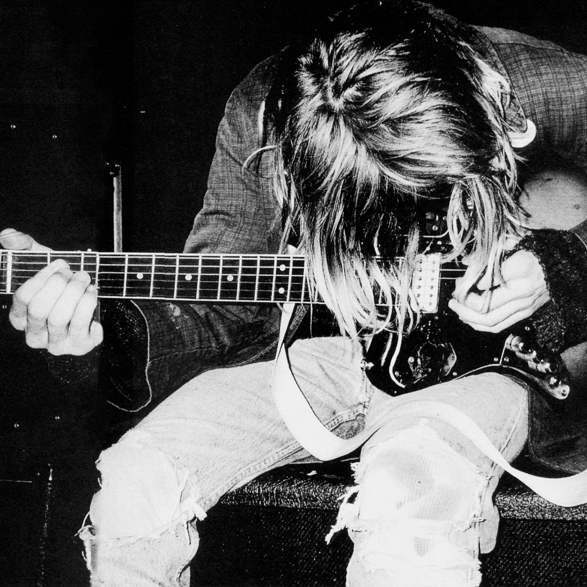 Cobain 桌布 壁紙 — 一張黑白照片，一位長髮音樂家坐著熱情地彈奏電吉他。