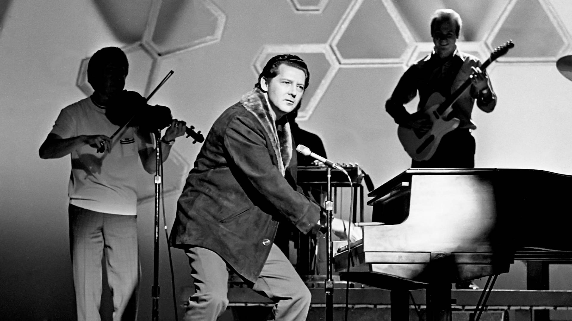 Jerry Lee Lewis 桌布 壁紙 — 一張黑白照片，一個樂團在舞台上表演，中央有鋼琴家，左邊有小提琴手，右邊有吉他手，背景是幾何圖案牆壁。