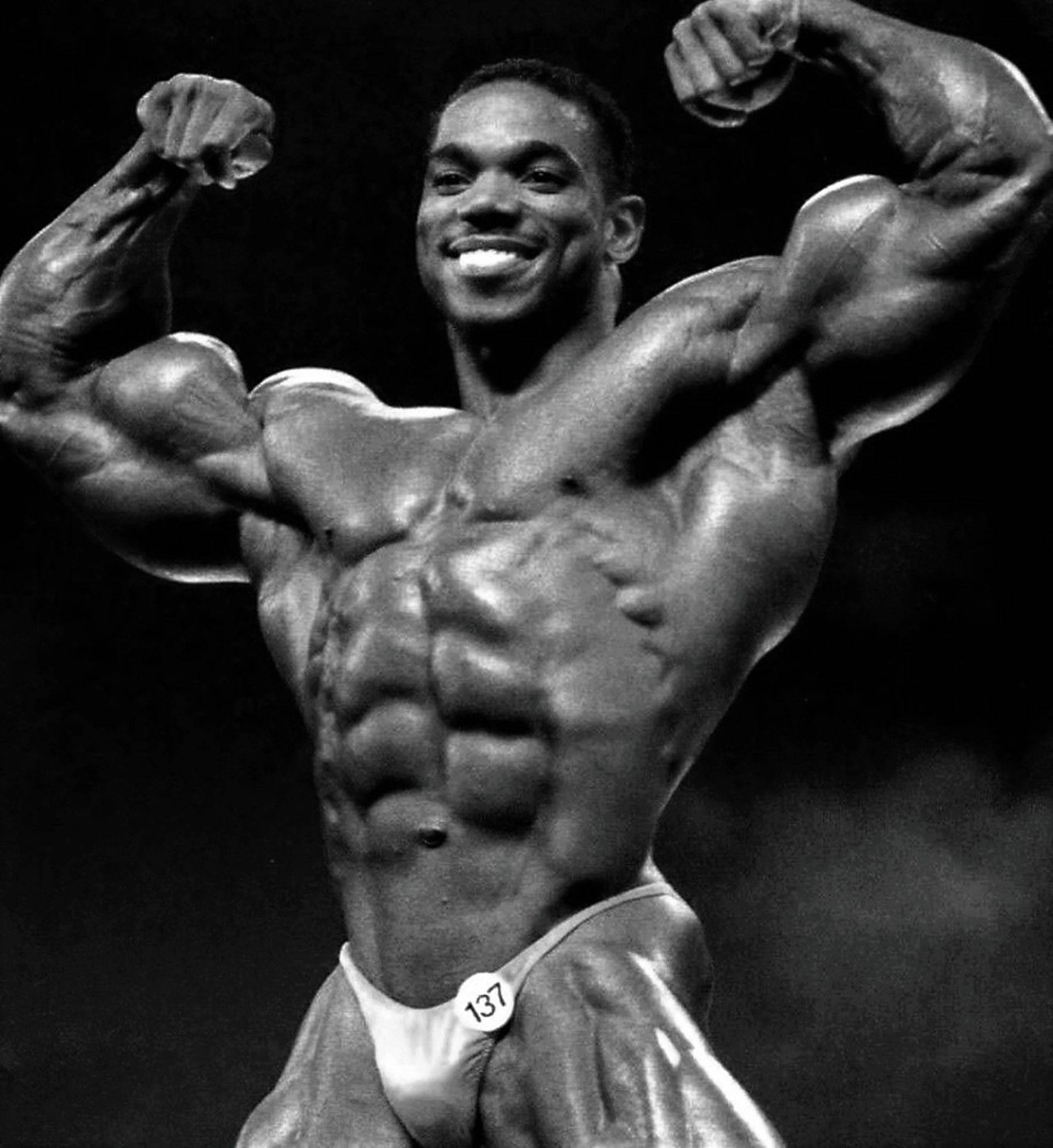 flex wheeler 桌布 壁紙 — 一張黑白照片，一名微笑的肌肉健美運動員在深色背景前彎曲雙臂。