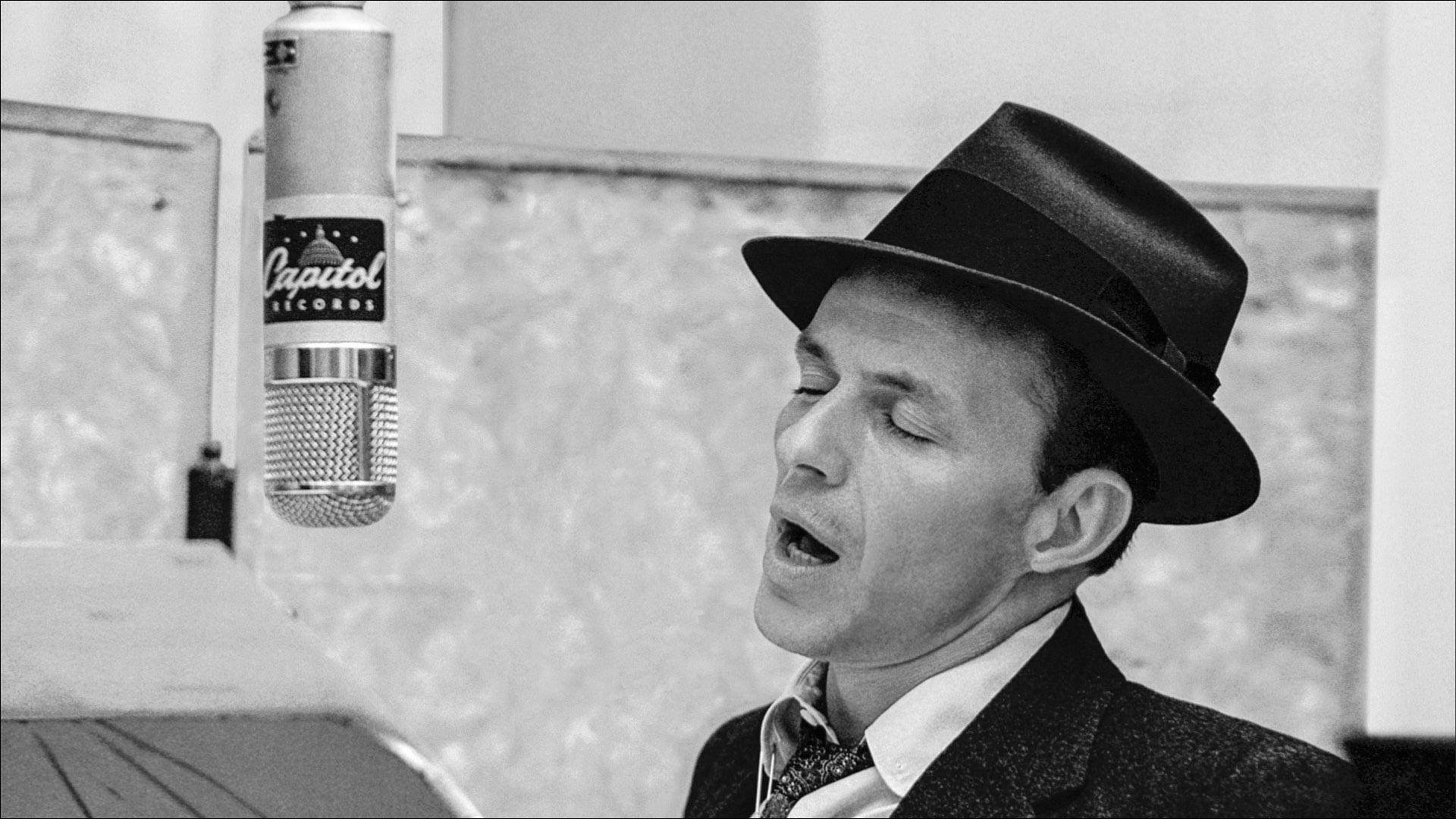 Sinatra 桌布 壁紙 — 一張黑白照片，一名戴著費多拉帽的男子在錄音室裡熱情地對著復古的Capitol Records麥克風唱歌。
