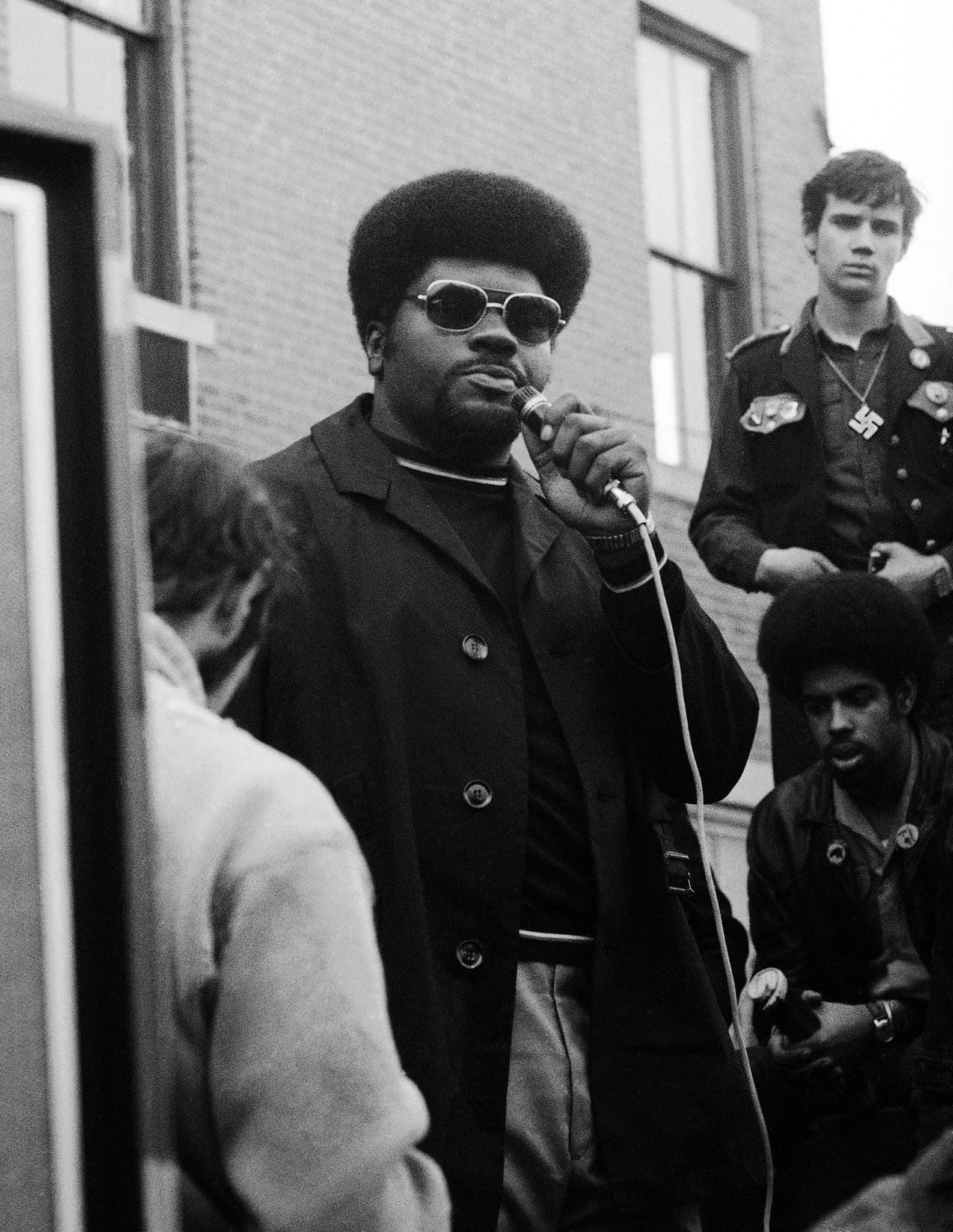 Black Panther Party 桌布 壁紙 — 一張黑白照片，一名留著非洲式髮型、戴著太陽眼鏡的男子在戶外對著麥克風說話。
