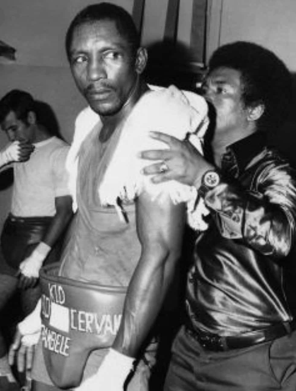 Boxing 桌布 壁紙 — 一張黑白照片，一名穿著印有「Kid Cervan」和「Tambabele」文字的短褲的拳擊手，正由一名穿著閃亮外套的男子照料。