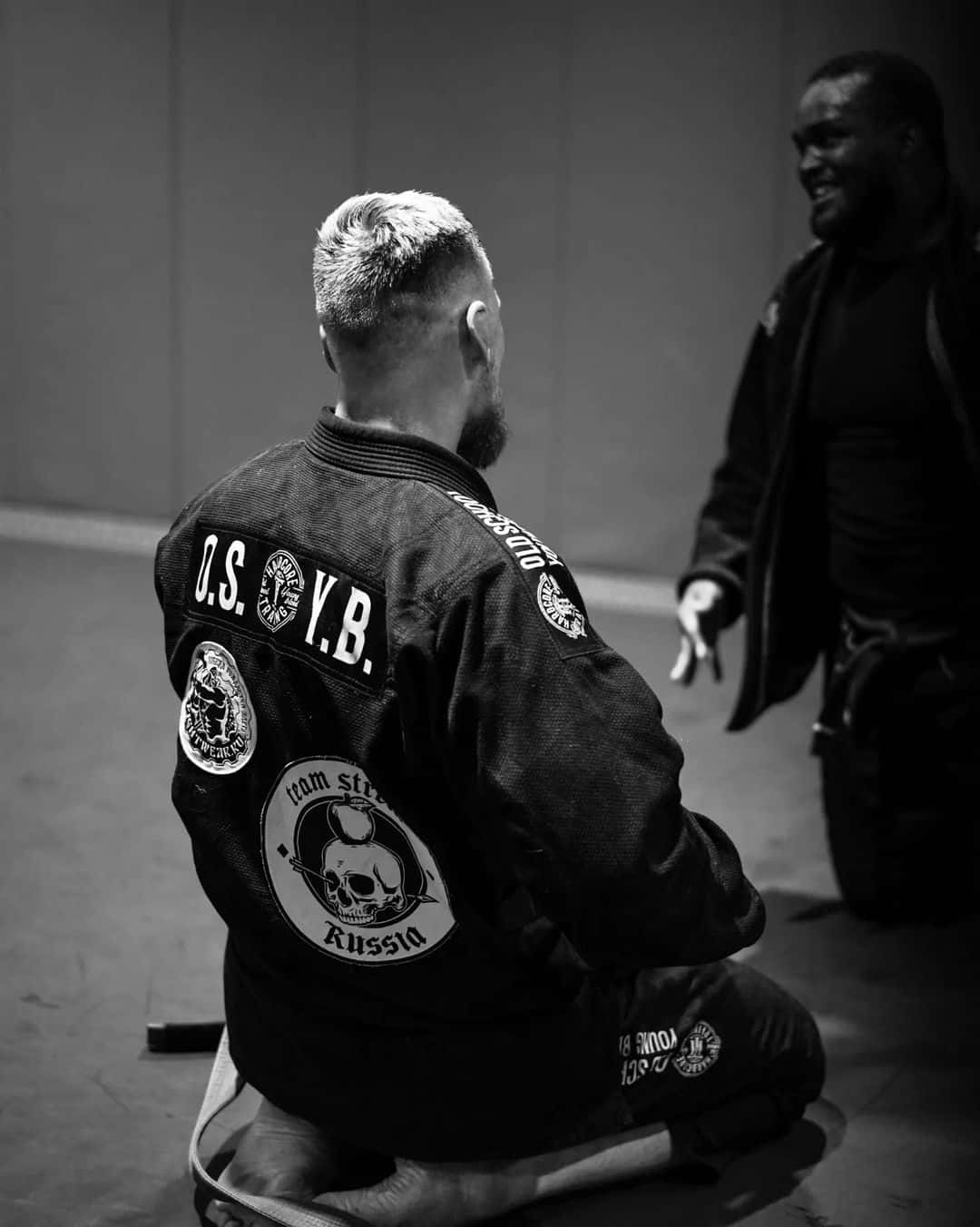 Athletic 桌布 壁紙 — 一張黑白照片，一名穿著武術道服的男子跪著，面向另一名男子，在燈光昏暗的健身房中。
