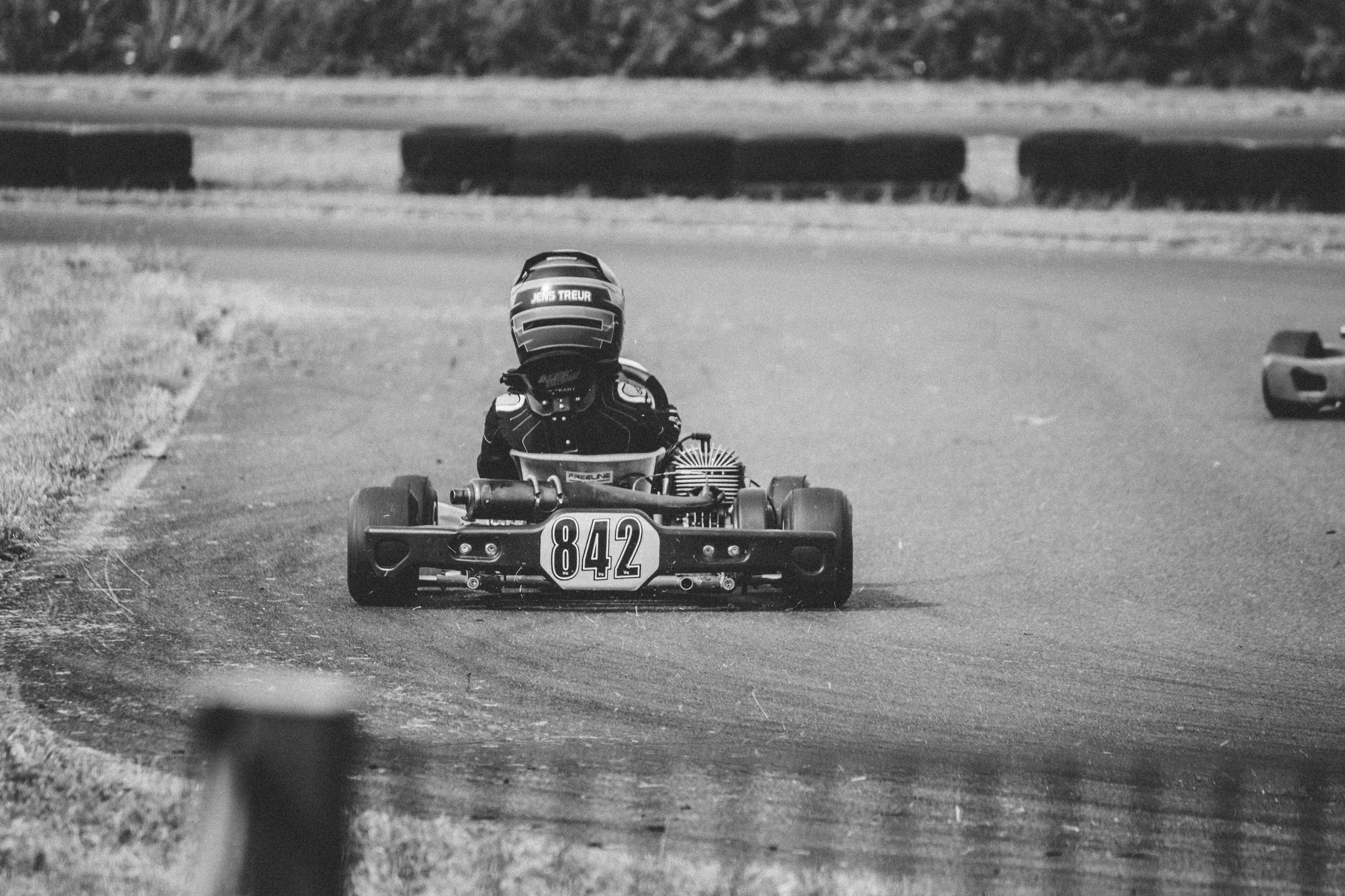 Racing 桌布 壁紙 — 一張黑白照片，一名身穿全套裝備的賽車手在賽道上疾馳，遠處可見另一輛賽車。