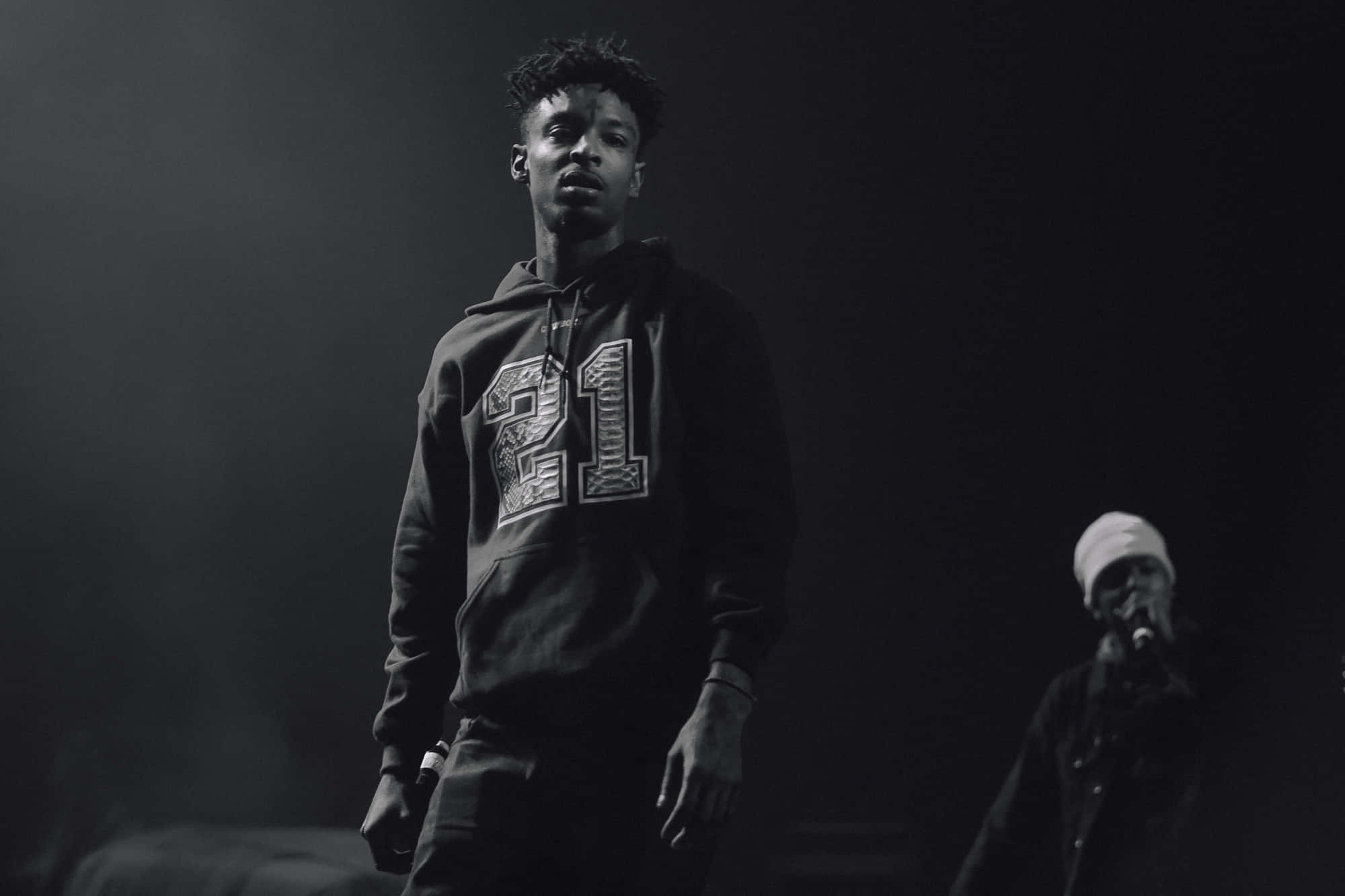 21 Savage 桌布 壁紙 — 一張黑白照片，一名身穿印有「21」連帽衫的年輕男子在表演期間站在燈光昏暗的舞台上。