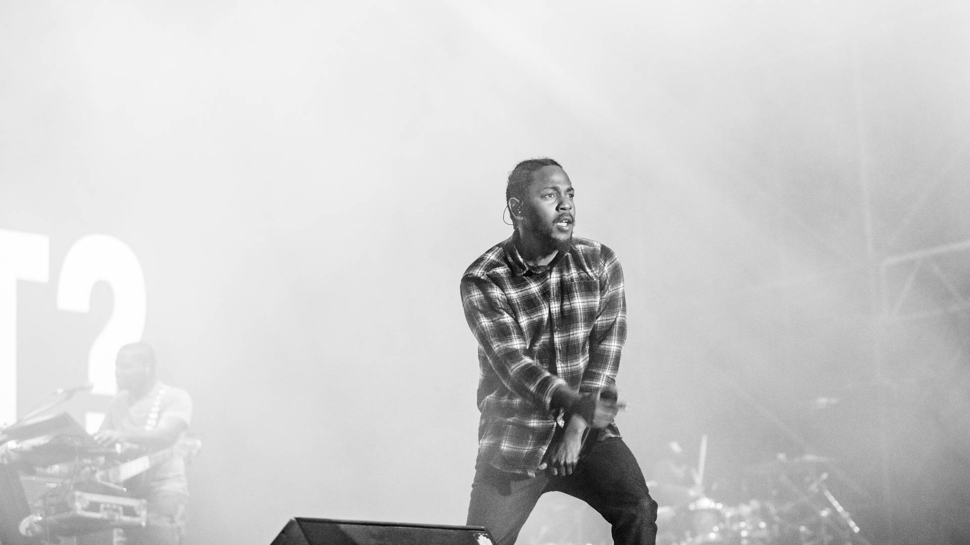 Kendrick 桌布 壁紙 — 一張黑白照片，一名身穿格紋襯衫的男表演者在煙霧和舞台燈光中蹲在舞台上。