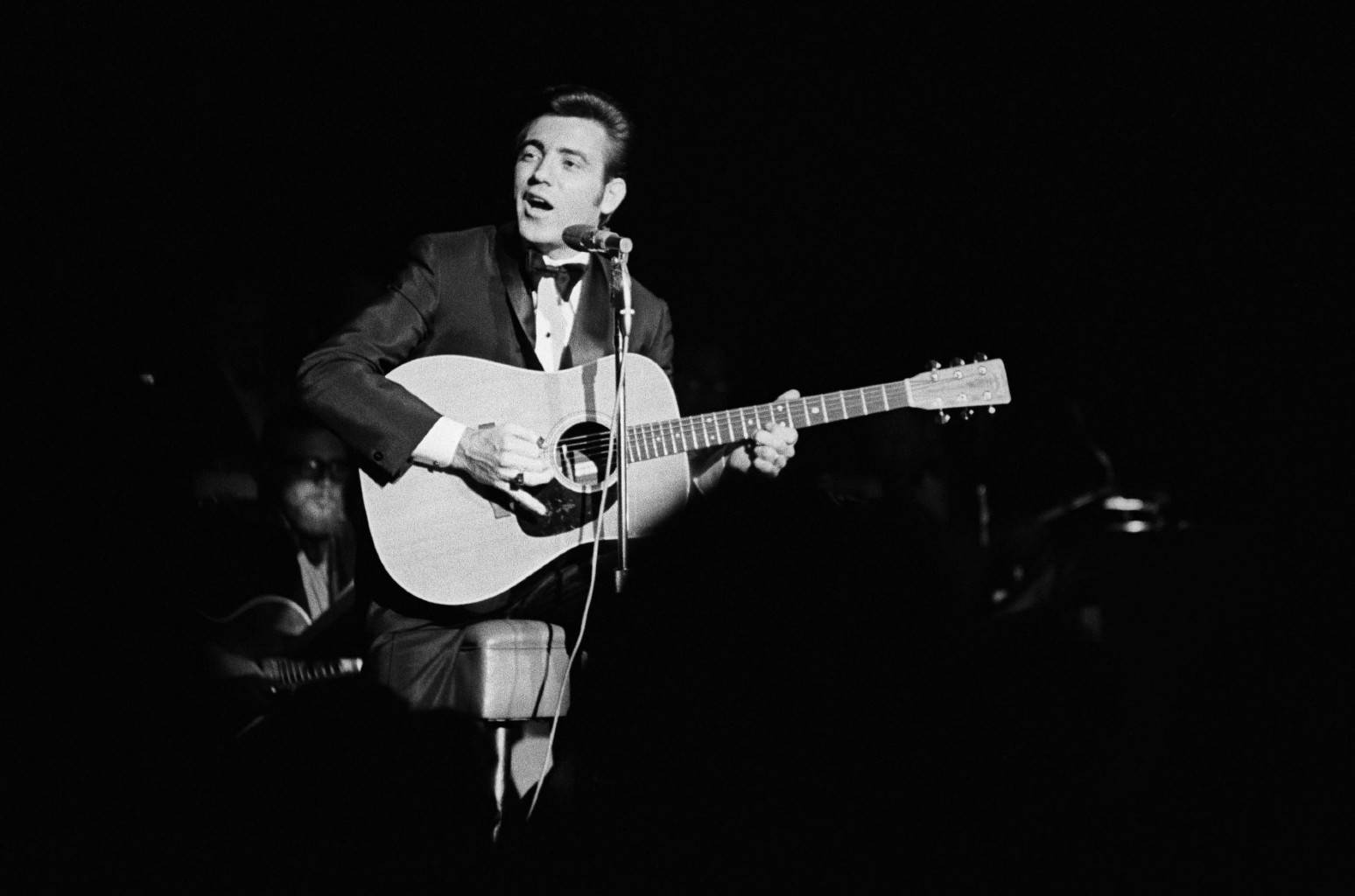guitar player 桌布 壁紙 — 一張黑白照片，一名身穿西裝的男子在舞台上對著麥克風唱歌，同時彈奏木吉他。