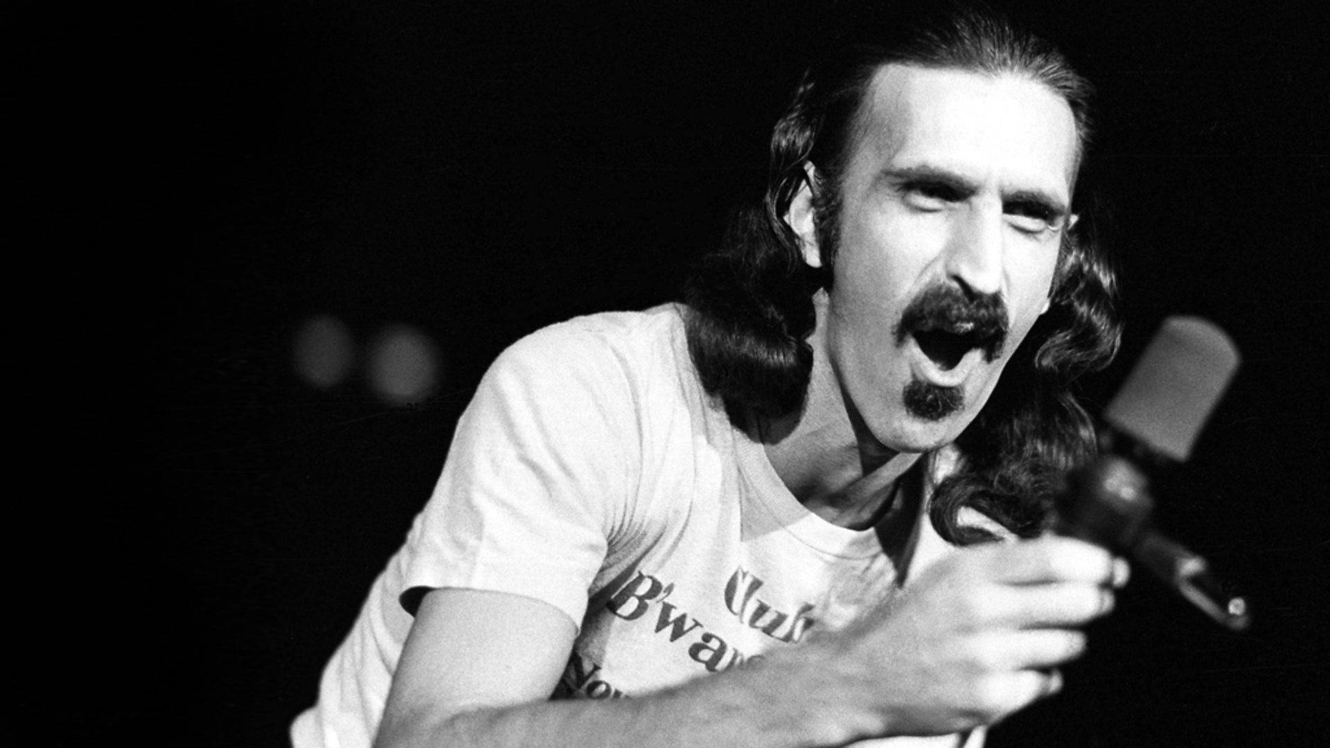 Zappa 桌布 壁紙 — 一張黑白照片，一名長髮留鬍子的男子在舞台上熱情地對著麥克風唱歌。
