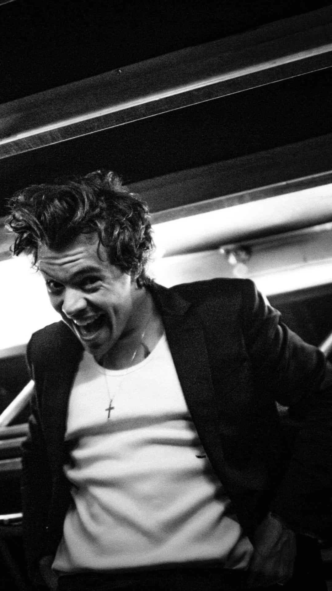 Harry Styles Black And White 桌布 壁紙 — 一張黑白照片，一名頭髮凌亂的男子在明亮的頭頂燈光下開懷大笑。