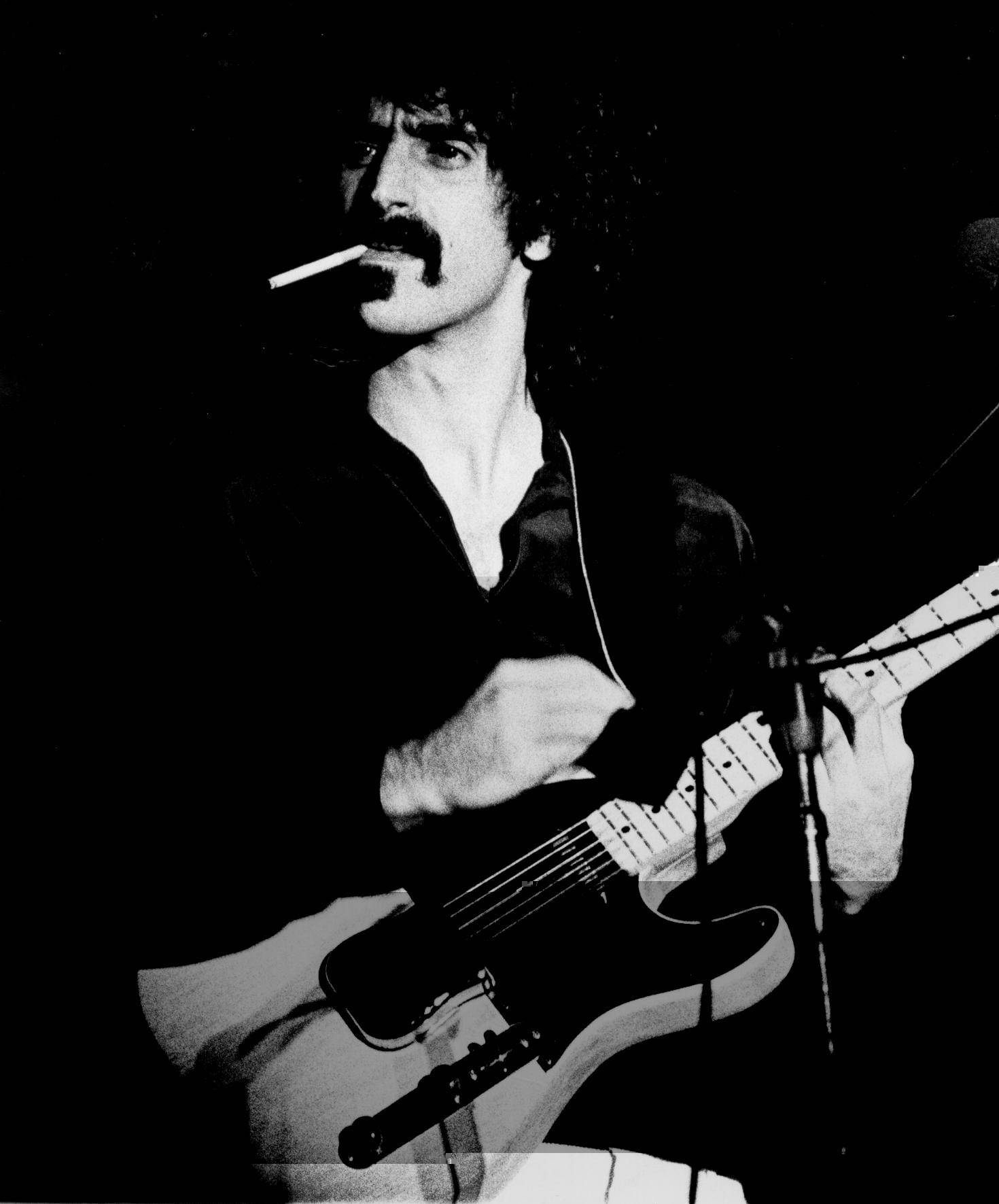 Zappa 桌布 壁紙 — 一張黑白照片，一名鬍鬚男嘴裡叼著煙，在舞台上彈奏電吉他。