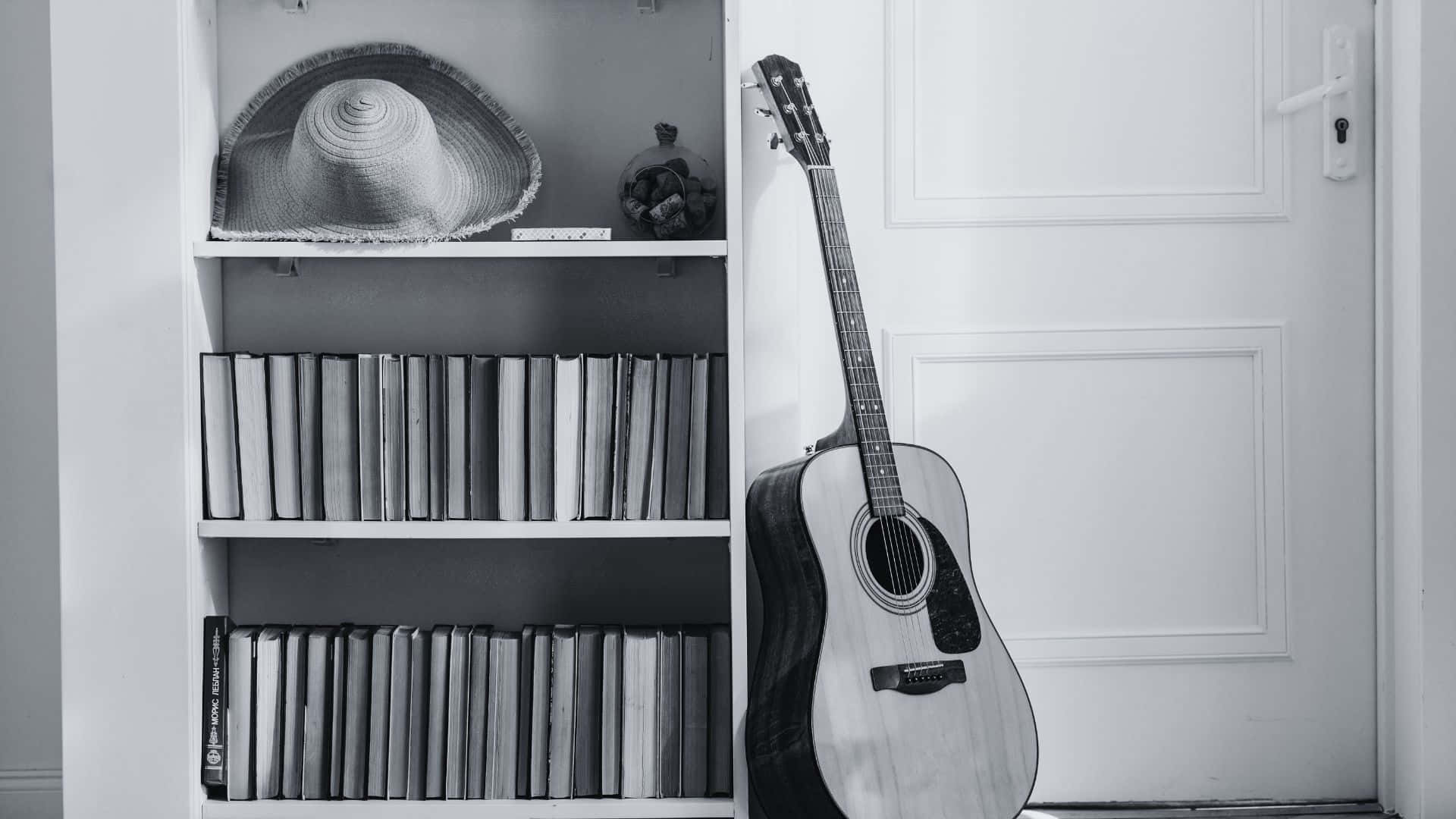 Black And White Guitar 桌布 壁紙 — 一張黑白照片，一把原聲吉他靠在一個裝滿書籍和草帽的書架旁邊。