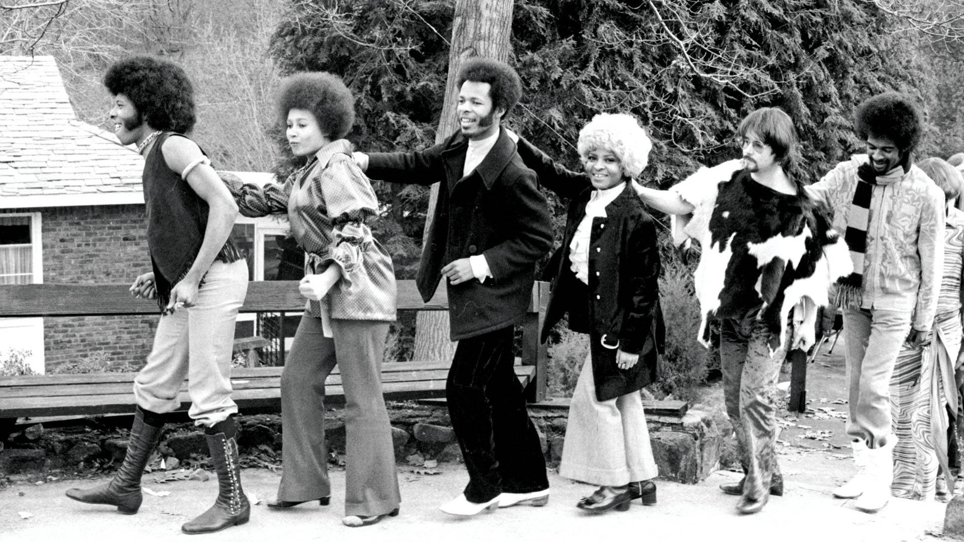 band 桌布 壁紙 — 一張黑白照片，六個身穿1970年代時尚的人士在戶外擺姿勢，背景有樹木和房屋。