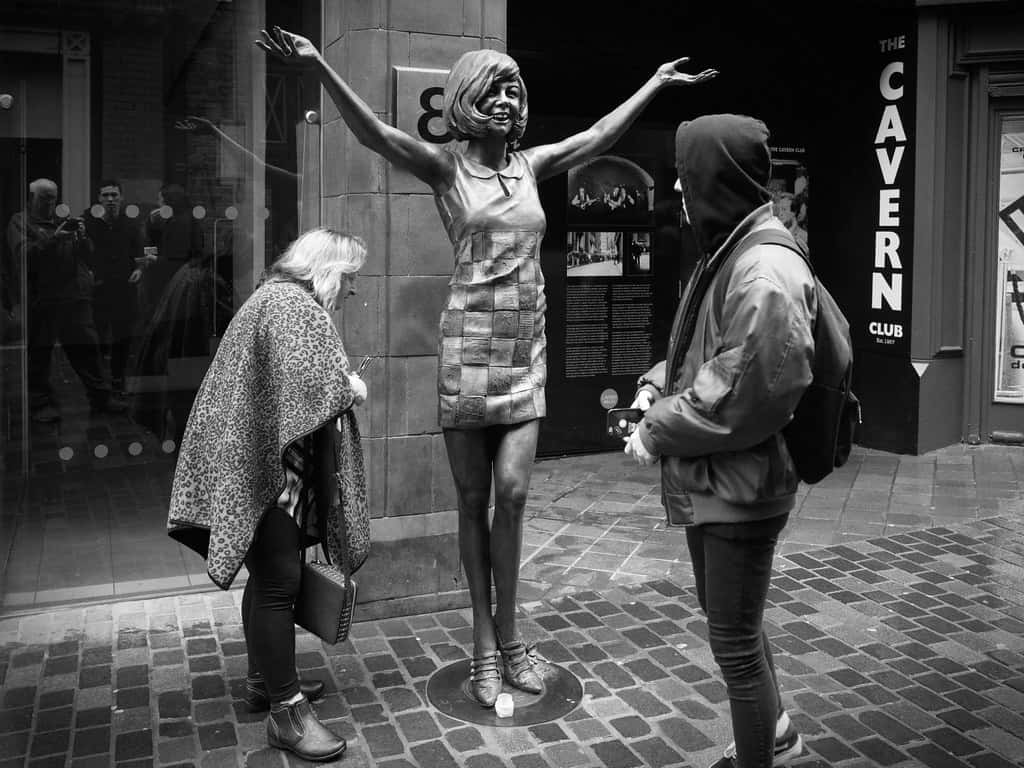 Statue Aesthetic 桌布 壁紙 — 一張黑白照片，展示一個真人大小、雙臂高舉的女性雕像，兩人在鵝卵石街道上觀察她。
