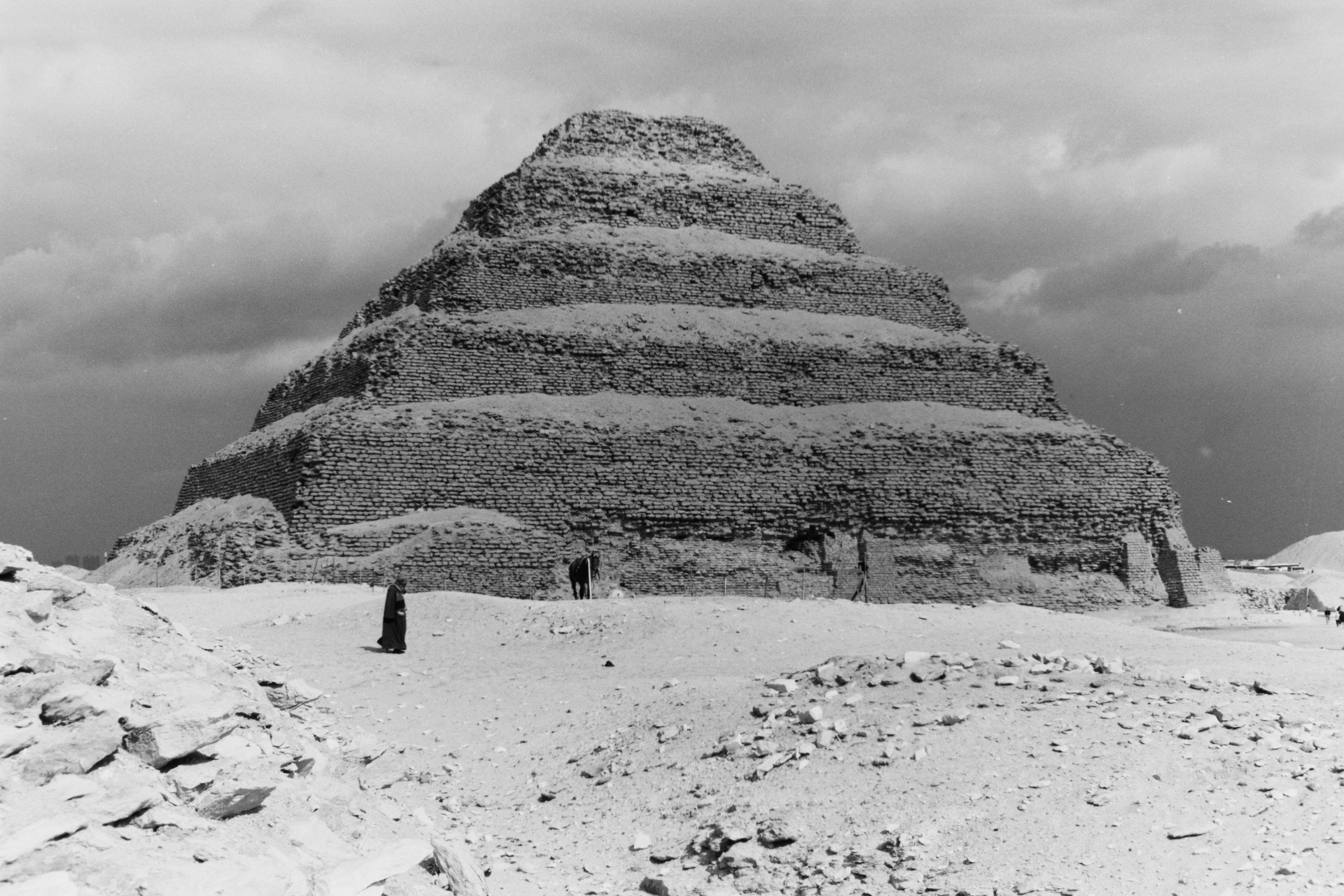 埃及 桌布 壁紙 — 一張黑白照片，展示左塞爾階梯金字塔在陰雲密佈的天空下巍然聳立，底部有微小的人影。
