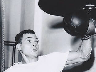 Boxing 桌布 壁紙 — 一張黑白照片，拍攝一位拳擊手在燈光昏暗的健身房中，對著沙包進行激烈訓練。