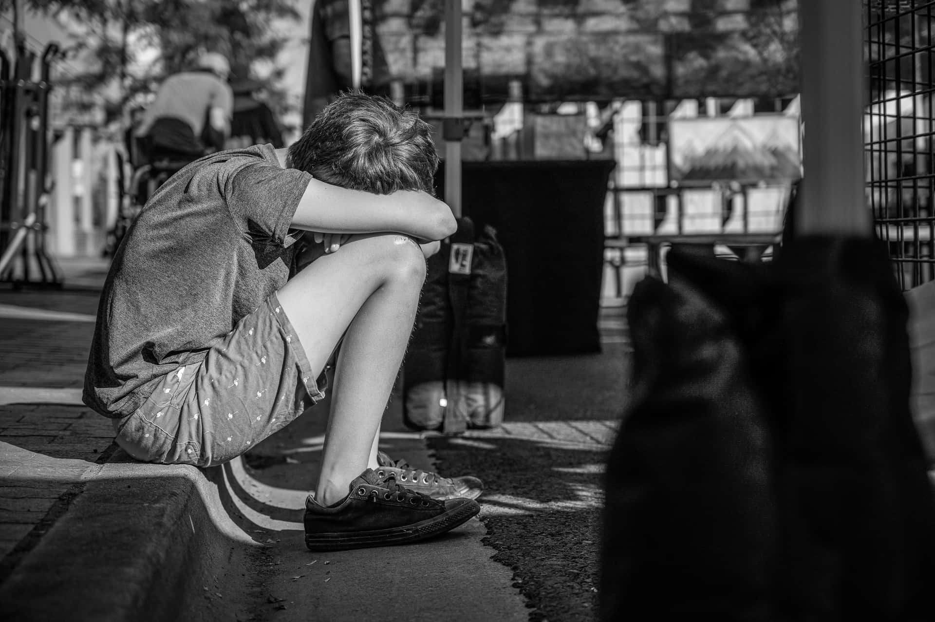 黑白 桌布 壁紙 — 一張黑白照片，拍攝一個孩子在城市環境中蜷縮在路邊，頭埋在膝蓋裡。