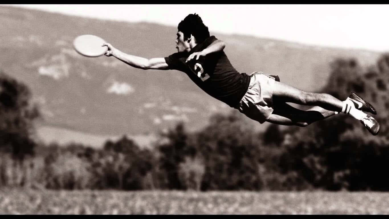 1366x768 Frisbee 桌布 壁紙 — 一張黑白照片，拍攝一名男子躍起接住飛盤，背景是朦朧的山巒。
